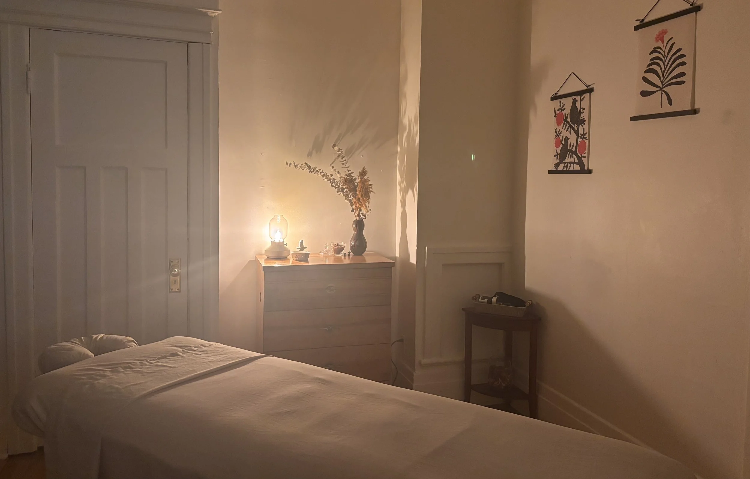 Une chambre chaleureuse avec un lit en premier plan et une lumière douce, une commode avec un vase de fleurs séchées, et des décorations murales avec des motifs floraux