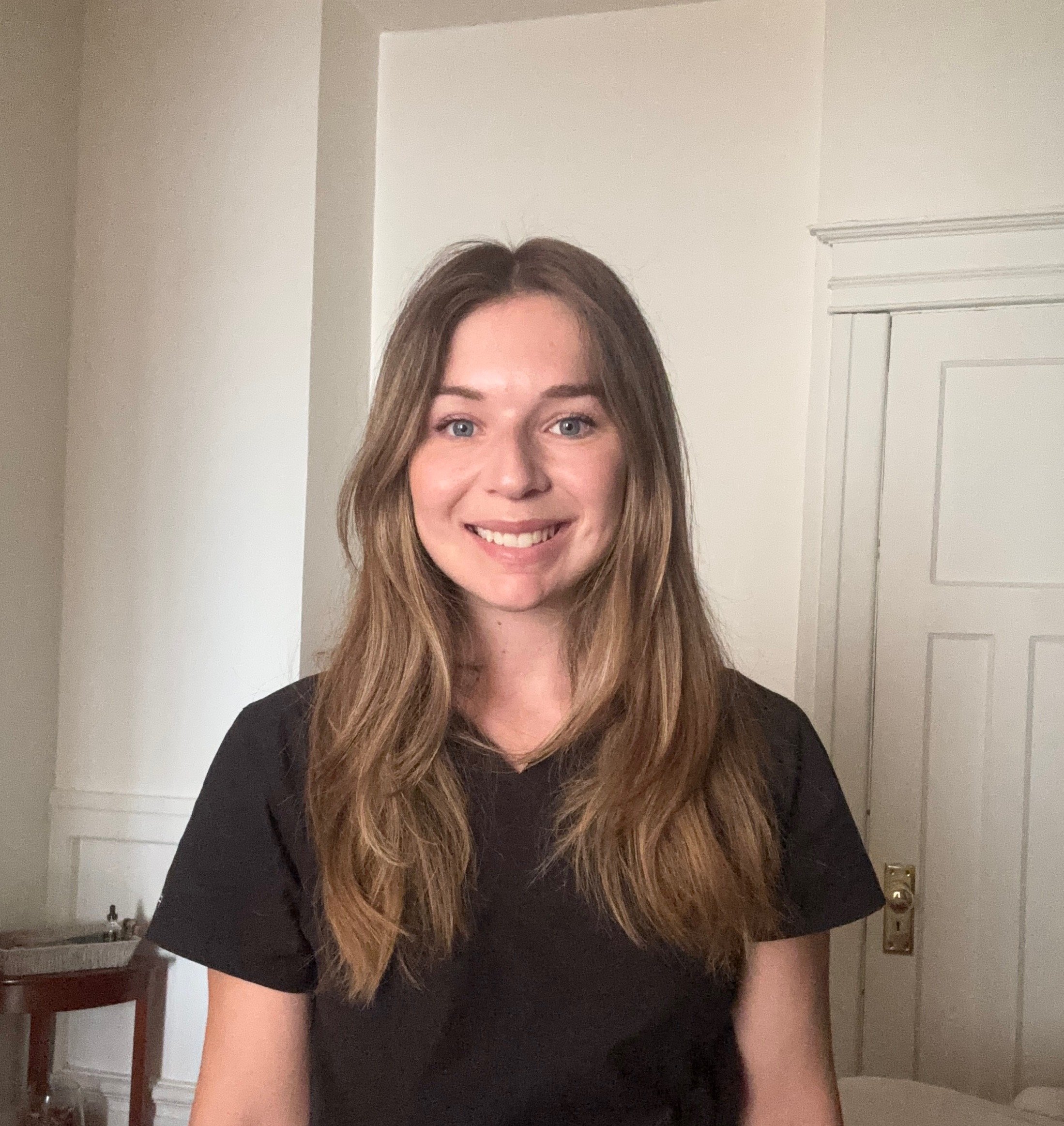 Jeanne, massothérapeute, souriante, portant un chandail noir, dans une pièce avec mur blanc.