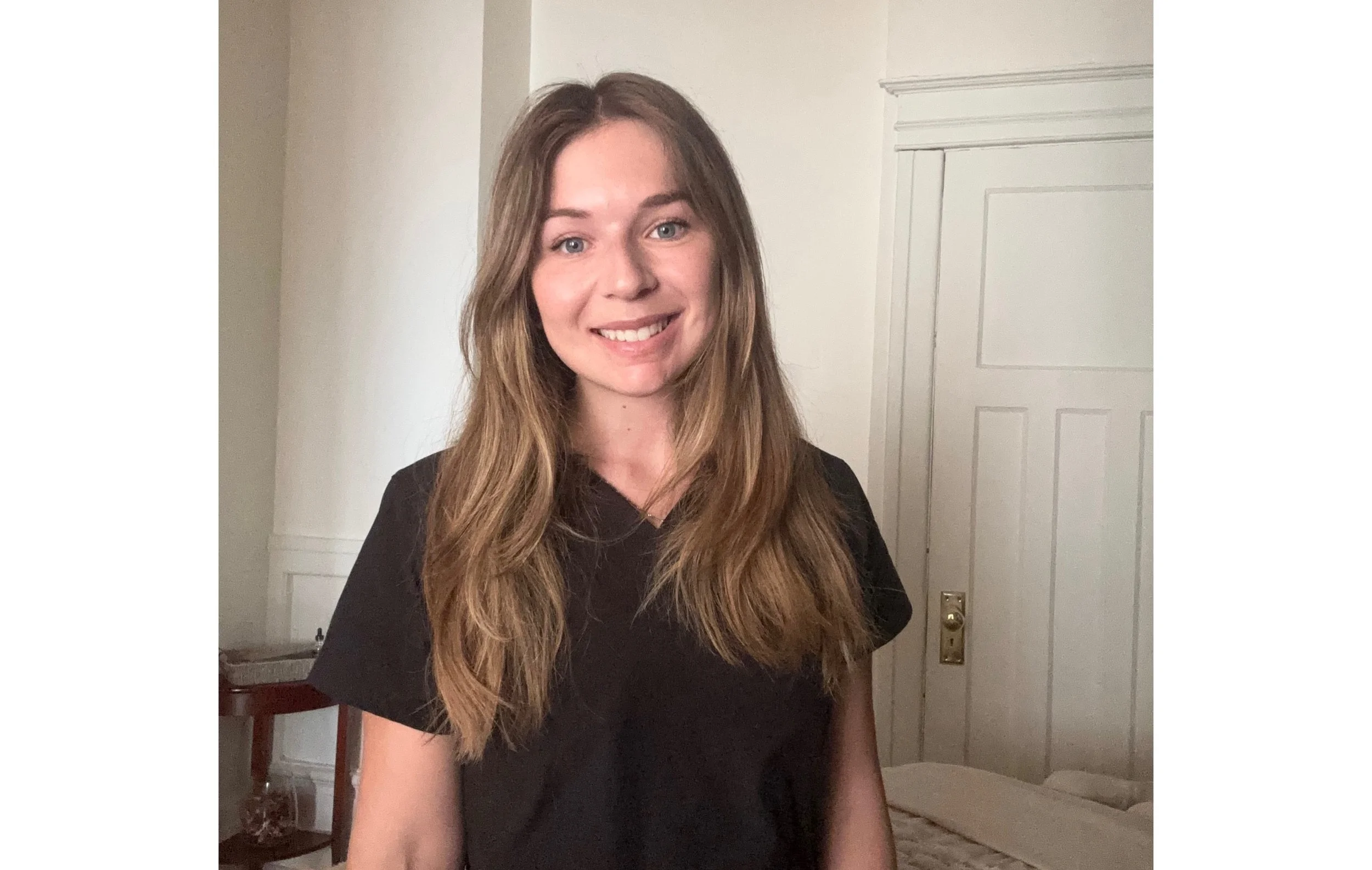 Jeanne, massothérapeute, souriante, portant un chandail noir, dans une pièce avec mur blanc.