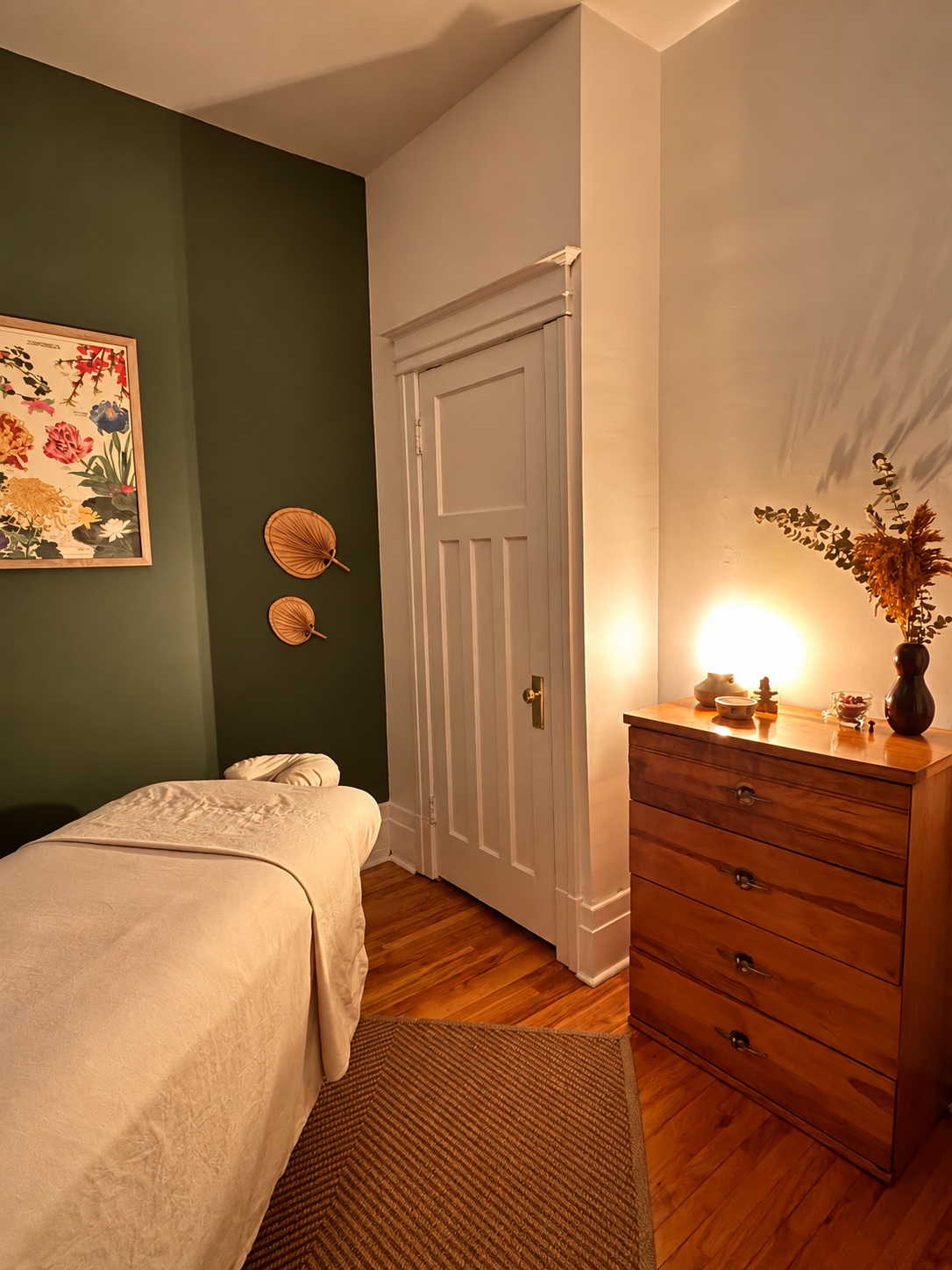 Studio de massothérapie à Rosemont montrant une pièce chaleureuse avec une table de massage, une commode en bois, une peinture florale, et une décoration avec des paniers en osier et des vases avec des plantes.