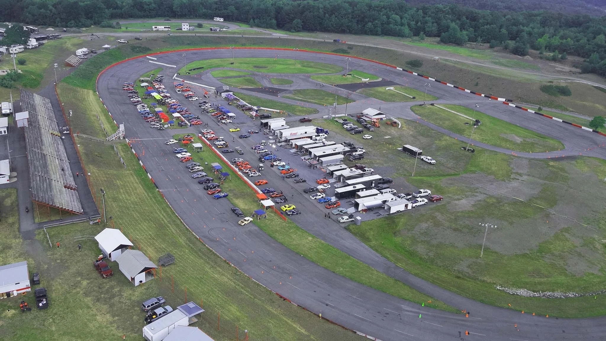 An aerial view of a go-kart racing track with parked cars and tents around it, surrounded by green hills and trees.