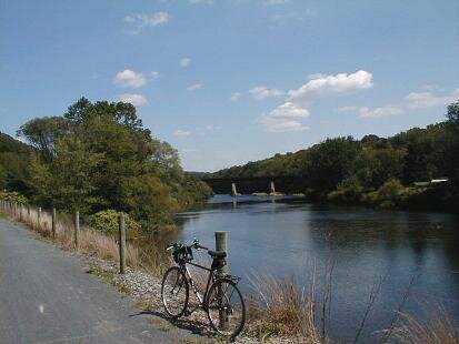 Roadside view with a bicycle parked near the water, trees on both sides, and a bridge in the background under a partly cloudy sky.