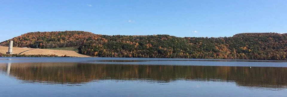 A calm lake reflecting a tree-covered hill with fall foliage, under a clear blue sky.