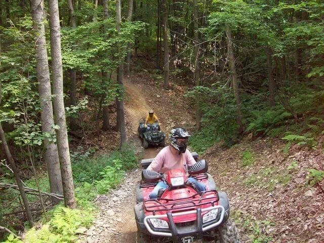 Two people riding all-terrain vehicles on a narrow, wooded dirt trail surrounded by trees and greenery.