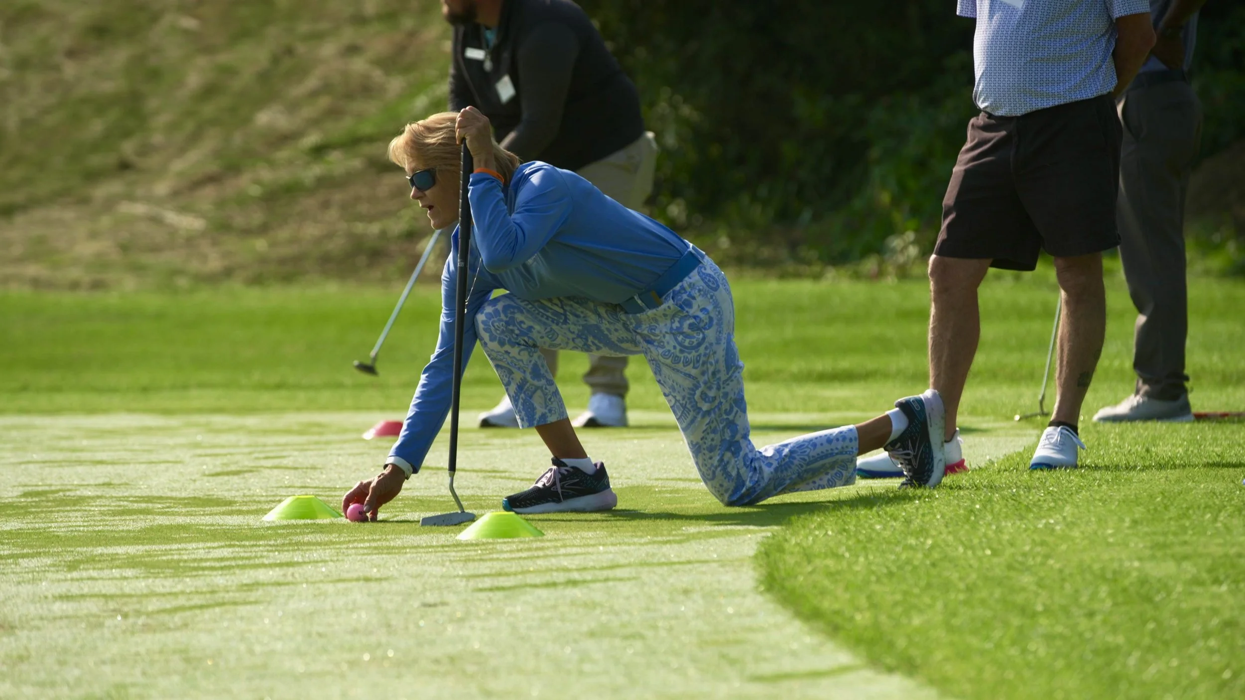 An elderly woman with sunglasses in a blue jacket and patterned pants is on her hands and knees putting a pink ball on the green putting green of a golf course, holding a putter in her left hand. Two men are standing nearby, observing her, with a woo