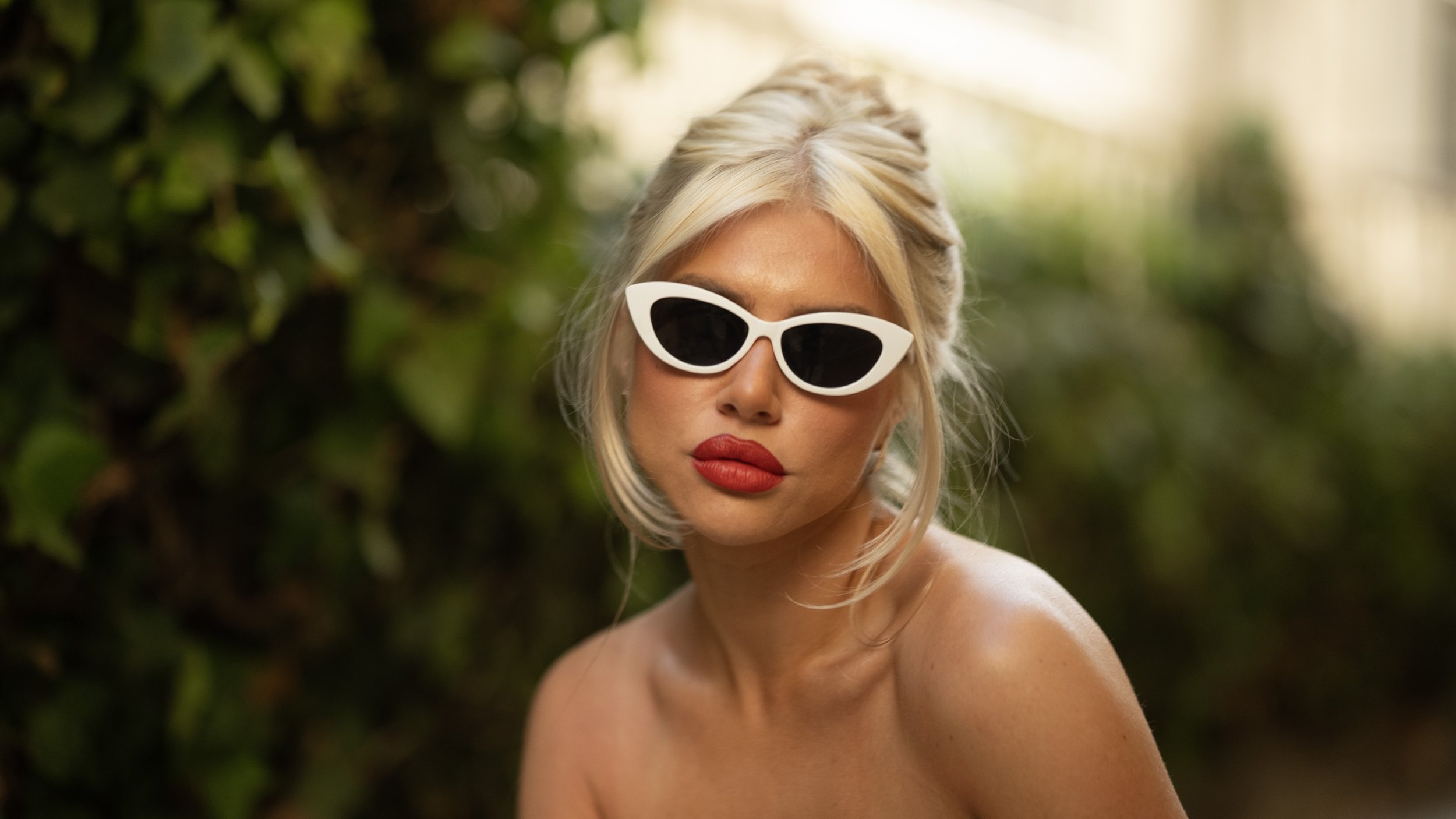 A woman with platinum blonde hair and red lipstick wearing white cat-eye sunglasses standing outdoors with green leafy background.