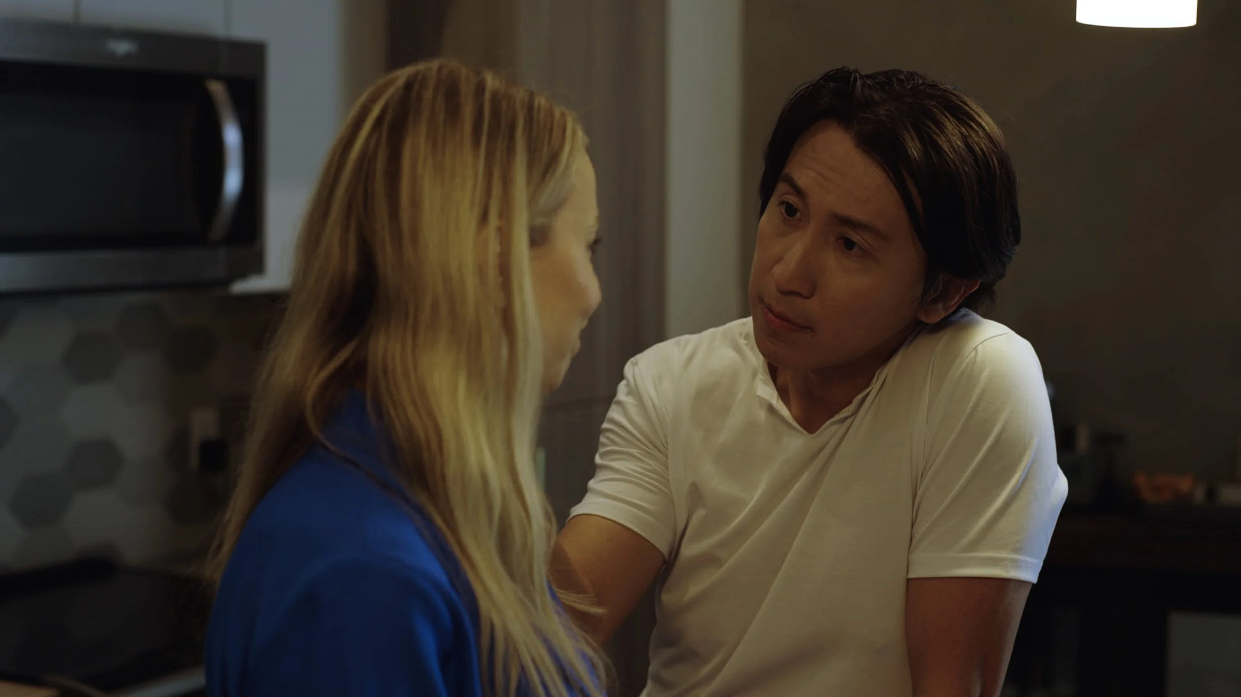 A man and a woman sit close together, facing each other, in a dimly lit room. The man has dark hair and is wearing a white t-shirt, while the woman has long blonde hair and is wearing a blue top. They appear to be engaged in a serious or intimate con