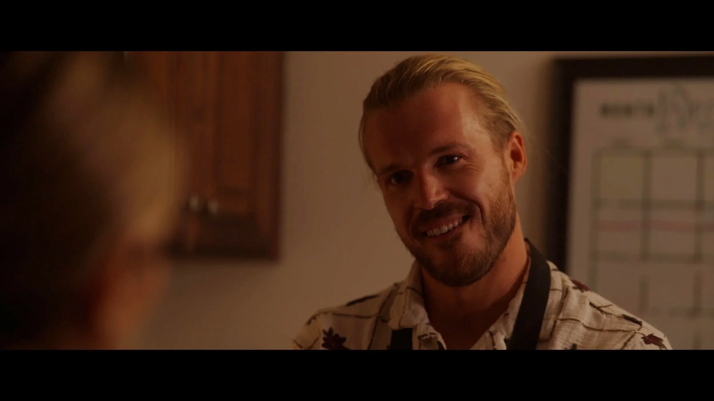 A man with a beard and blond hair smiling at another person in an indoor setting.