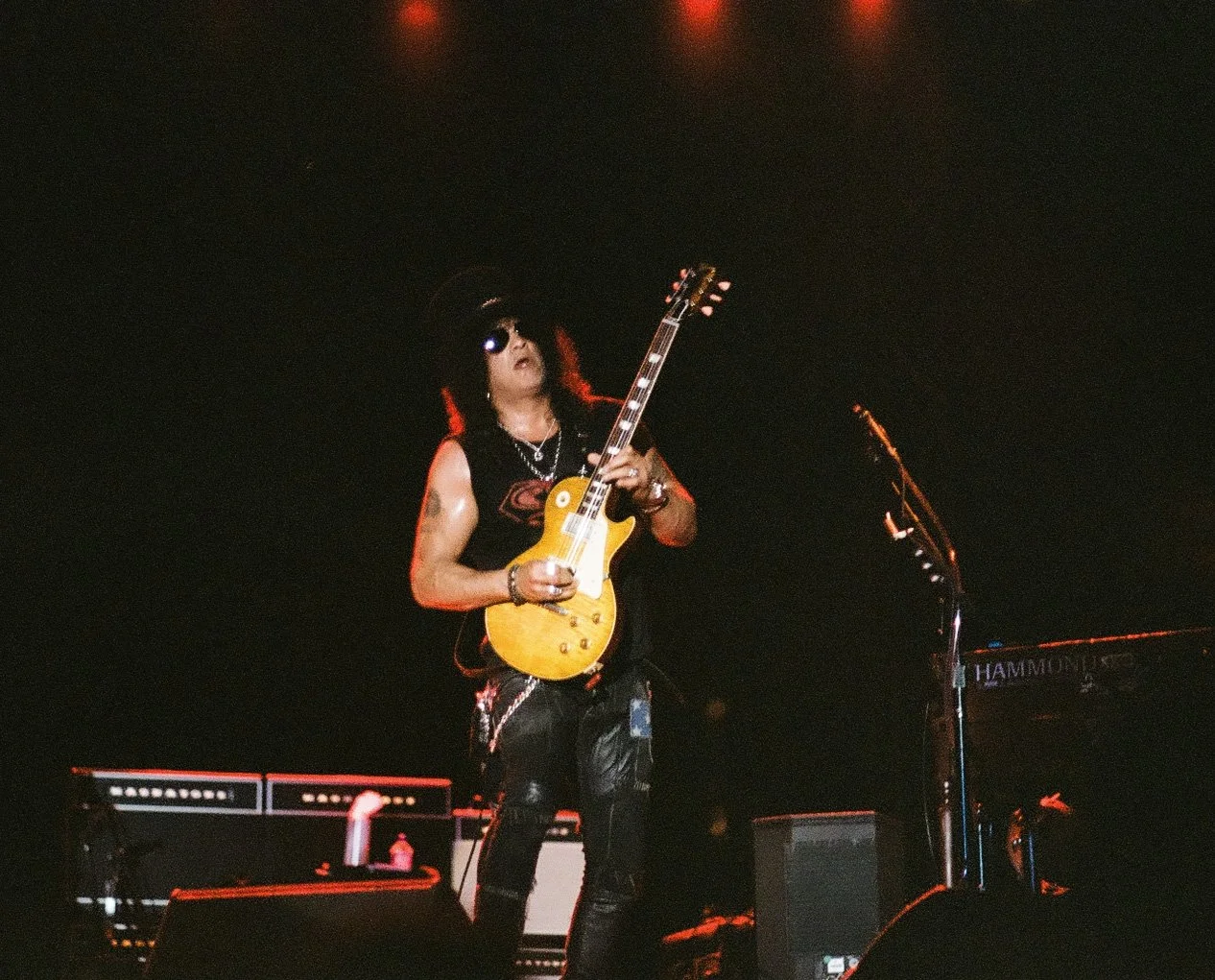 A musician playing a yellow electric guitar on stage, wearing sunglasses and a black tank top, with a dark background and stage equipment.