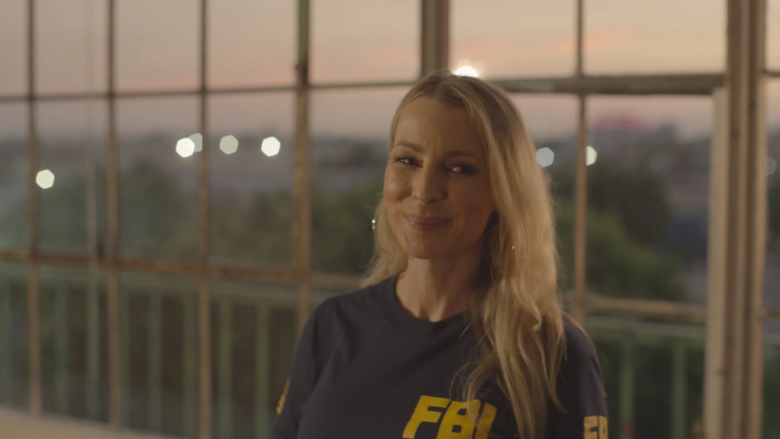 Smiling woman with long blonde hair standing in front of a window with a cityscape background at sunset.