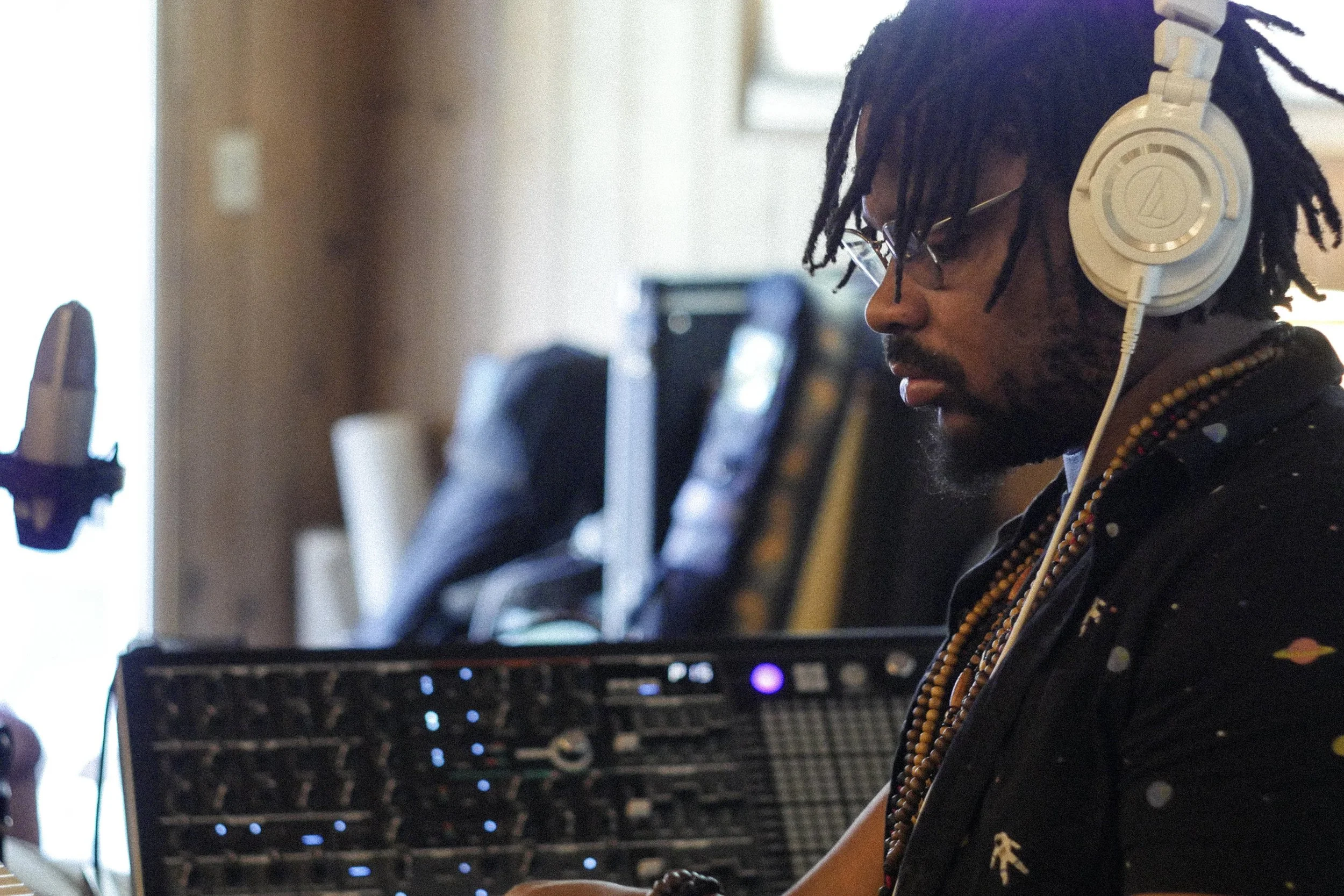 A man with dreadlocks and glasses, wearing headphones and a beaded necklace, working on a music mixer in a studio.