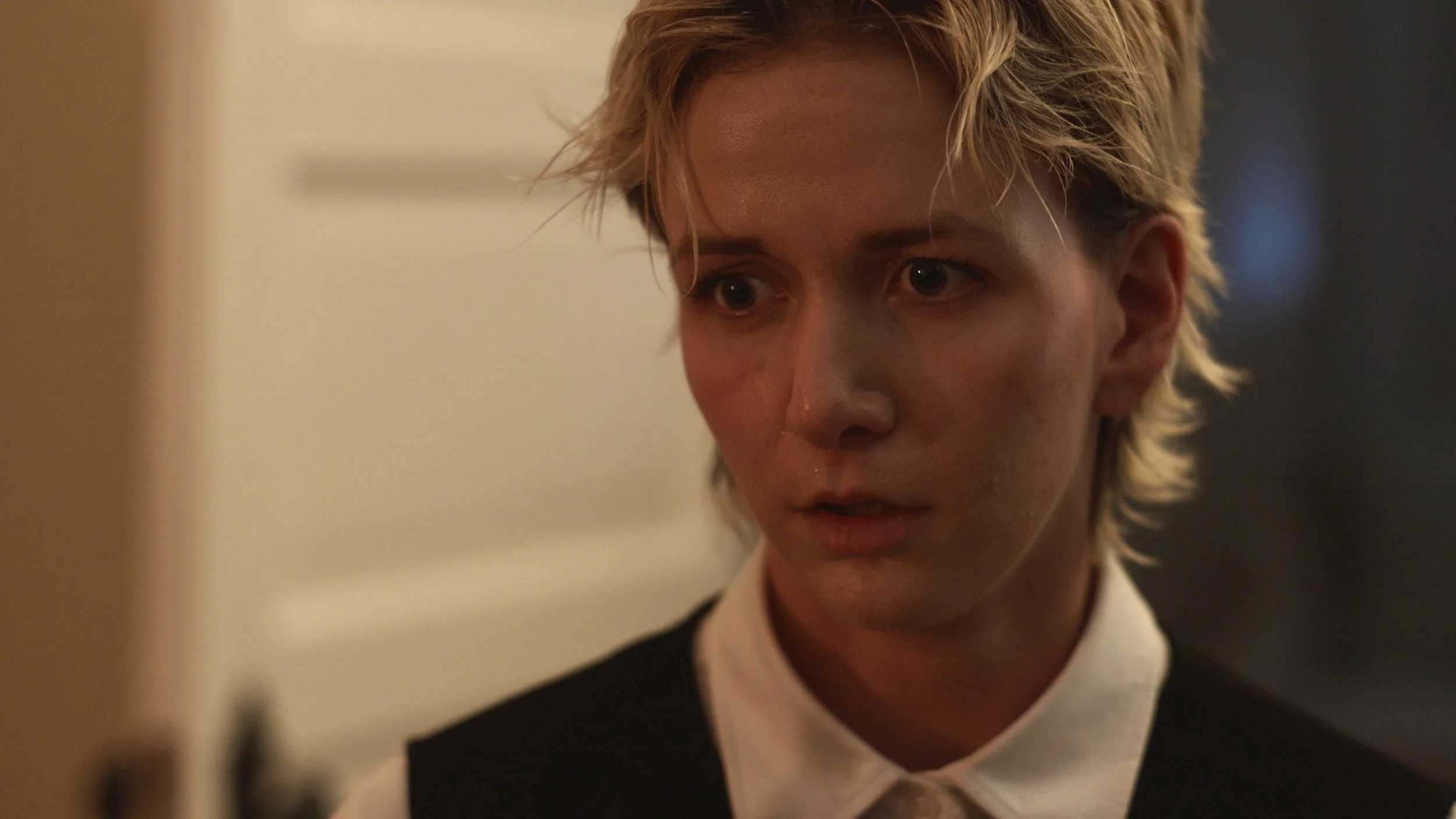 Close-up of a young woman with short blonde hair, wearing a white collared shirt and a black vest, looking thoughtful.