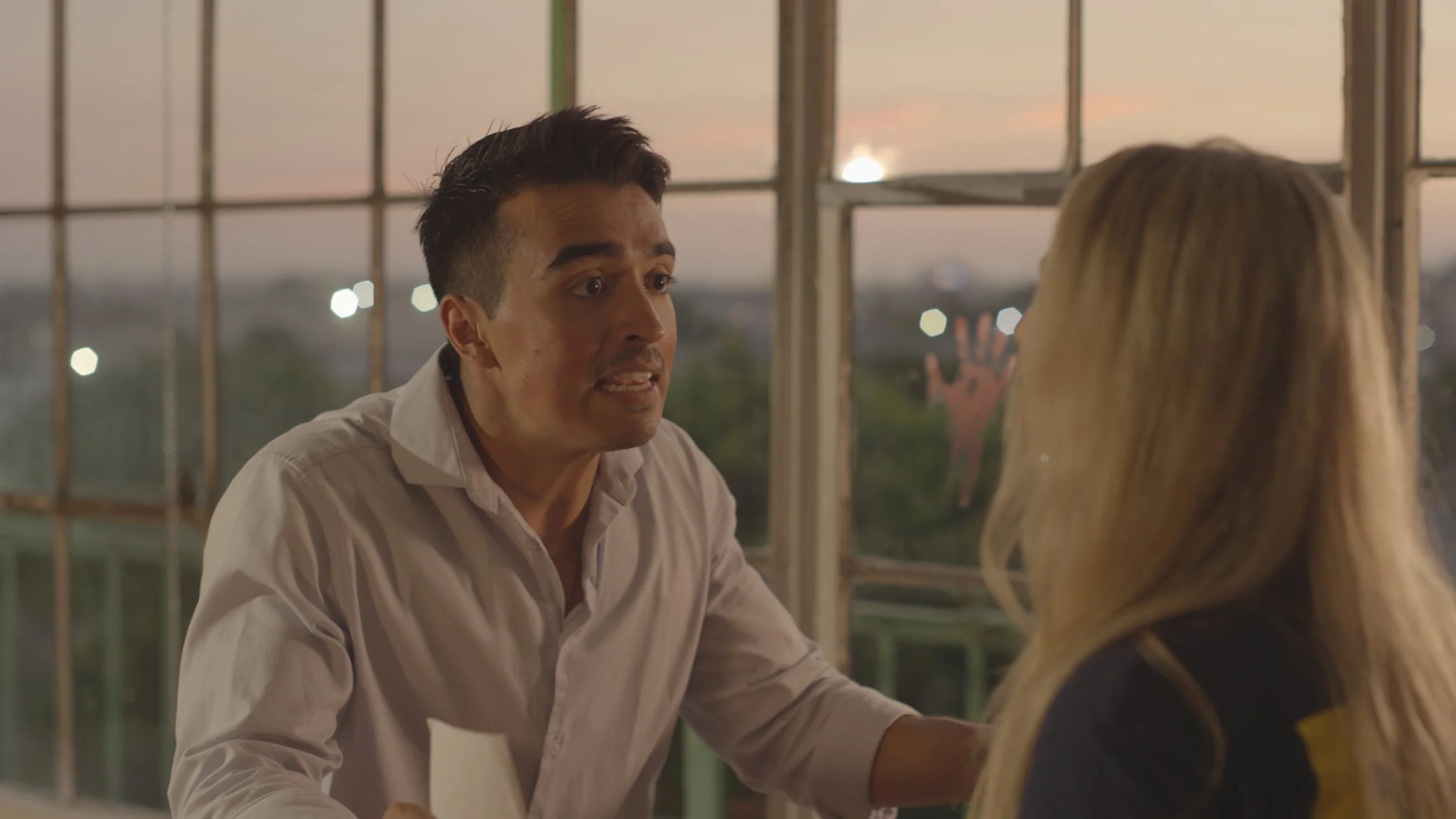 A man with dark hair wearing a white shirt is speaking to a woman with blonde hair. They are sitting at a table with large windows behind them, showing the sunset and city lights outside.