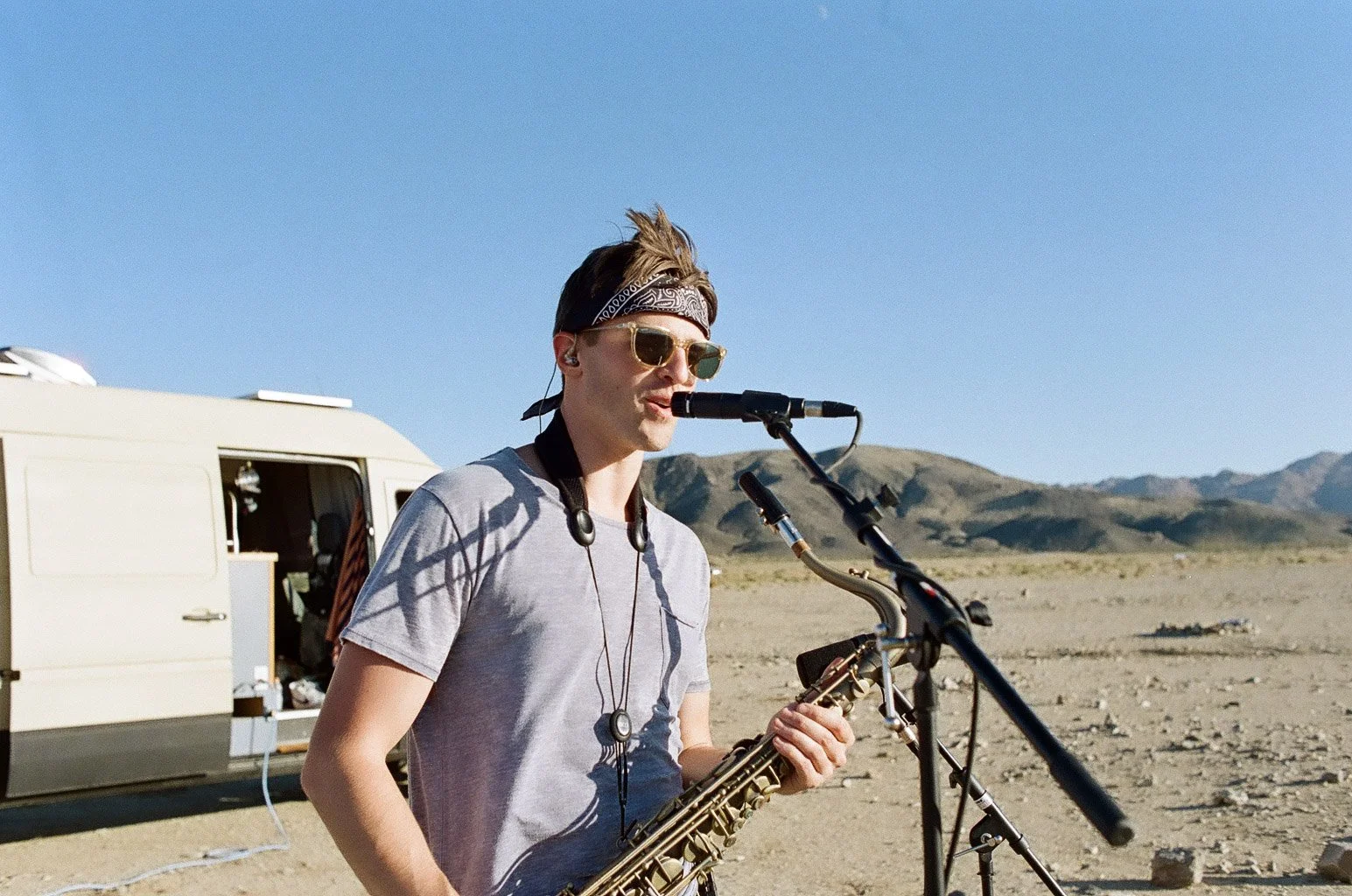 A young man wearing sunglasses, a bandana, and a gray t-shirt playing a saxophone outdoors with a desert landscape and mountains in the background. There is a trailer behind him.