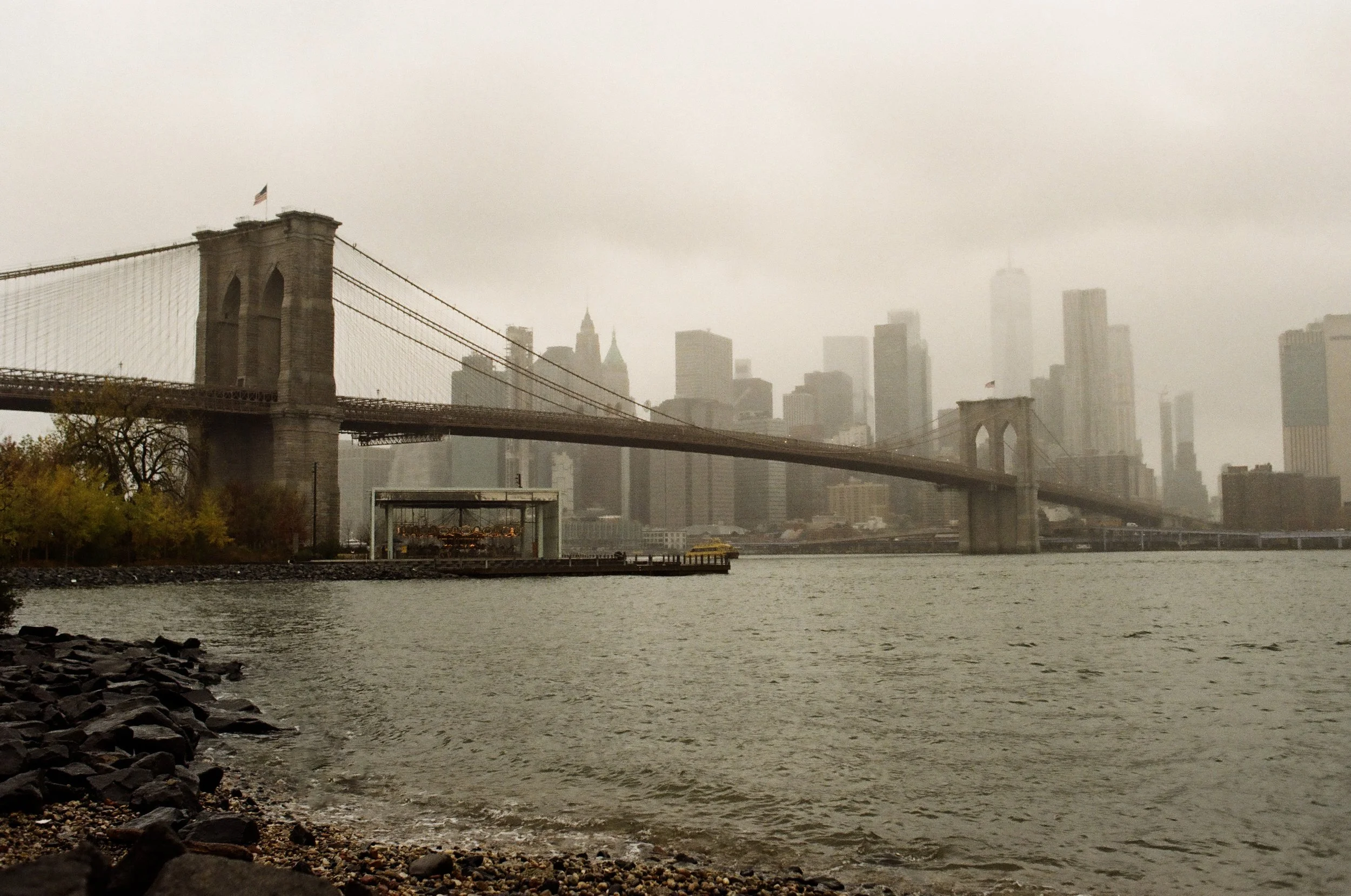 A view of the Brooklyn Bridge with New York City skyscrapers in the background on a cloudy, foggy day.