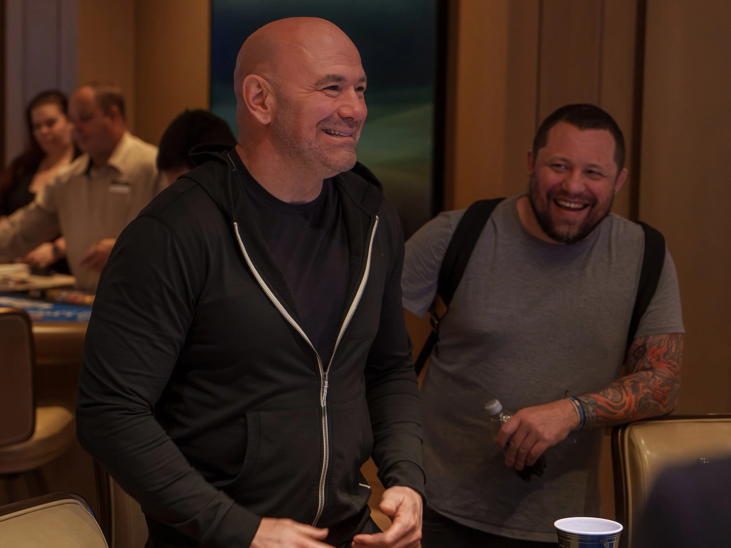 Two men are smiling and enjoying themselves at an indoor gathering. The man on the left is bald and wearing a black jacket, while the man on the right has short hair, a beard, tattoos on his arm, and is wearing a gray T-shirt. They are surrounded by 