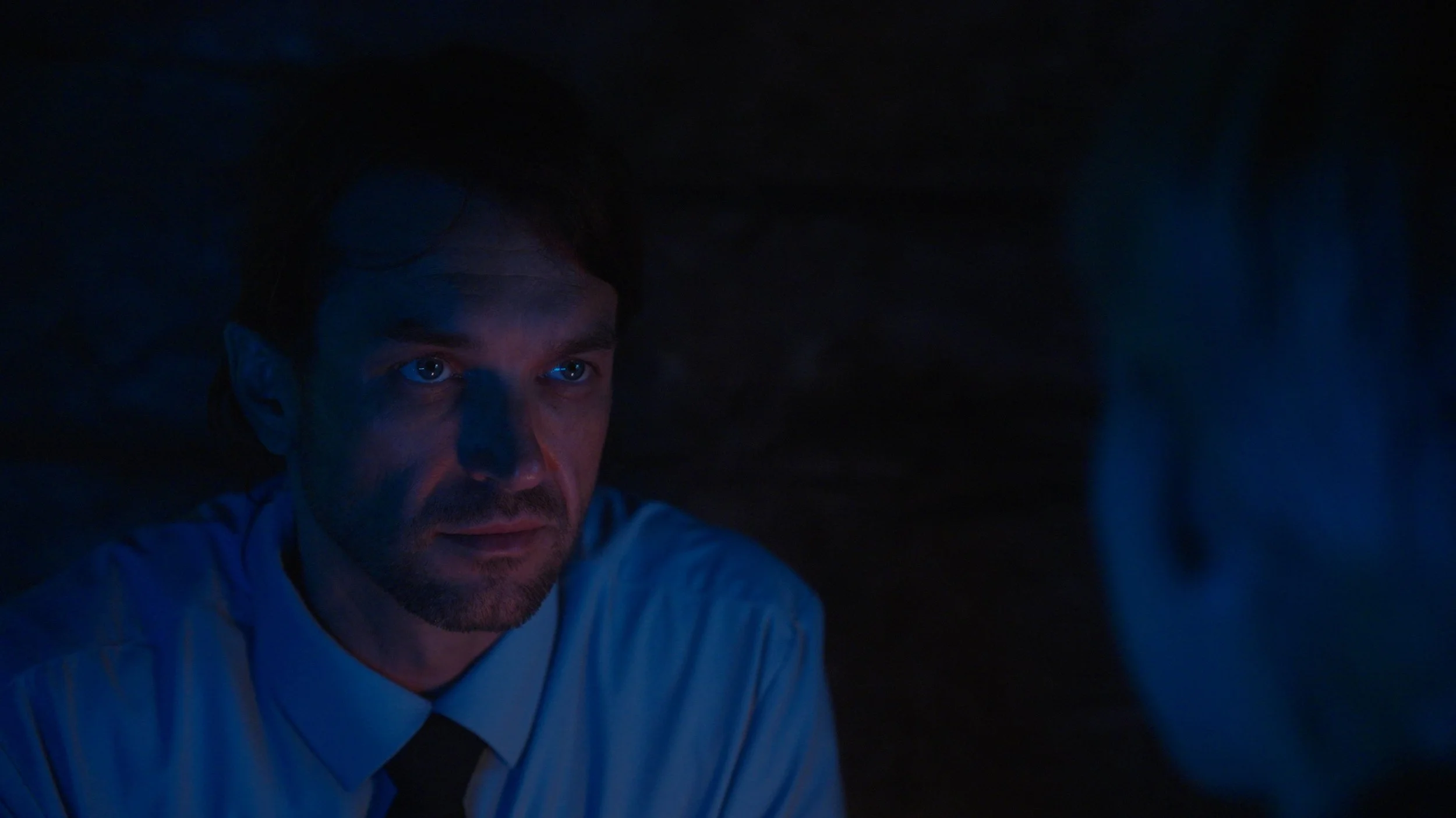 A man with blue eyes and dark hair looking intently in a dark room with blue lighting.
