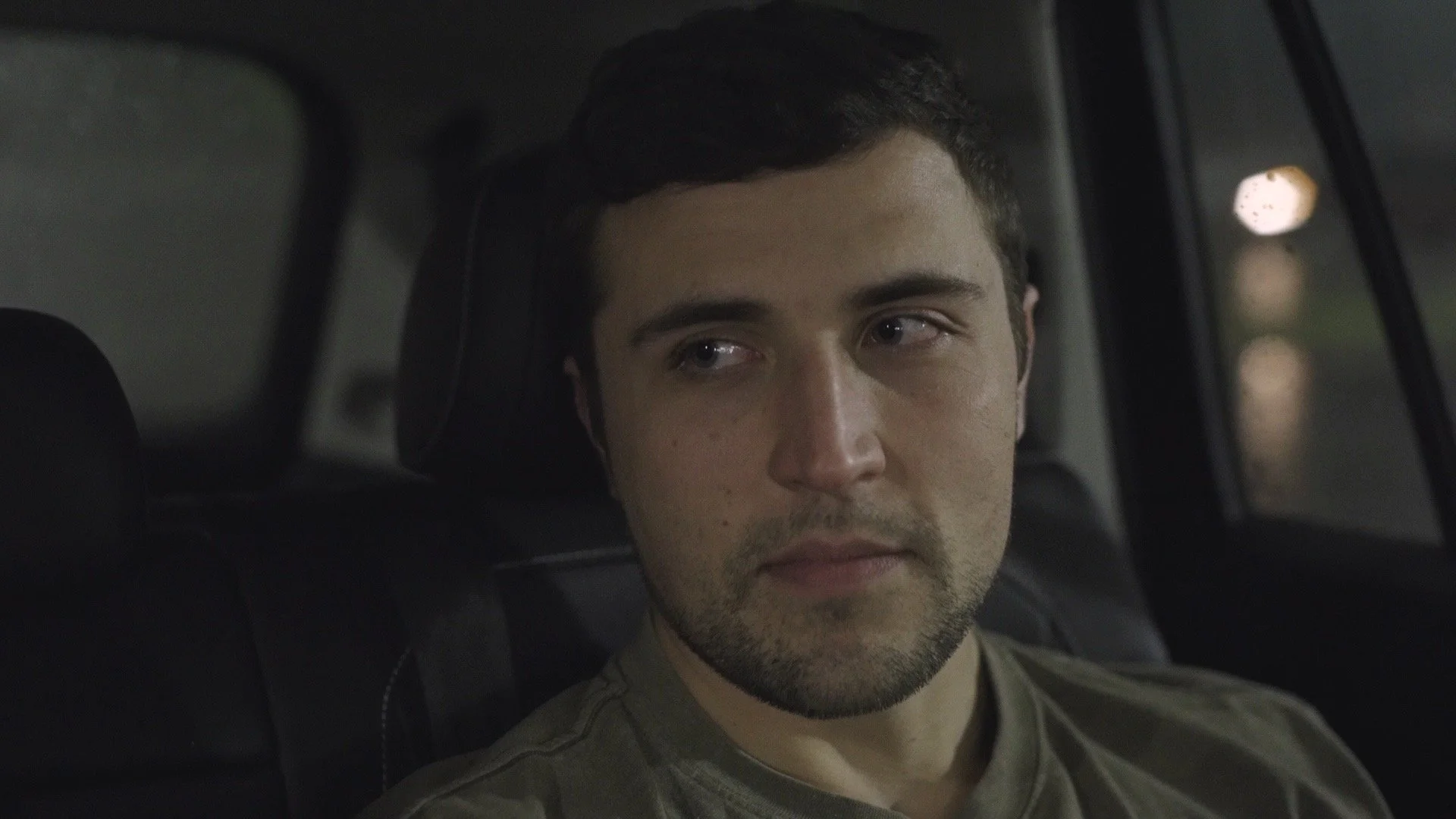 A young man with short dark hair and stubble is sitting in the backseat of a vehicle, looking to his right with a neutral expression. The background is blurry with faint lights visible outside the window.