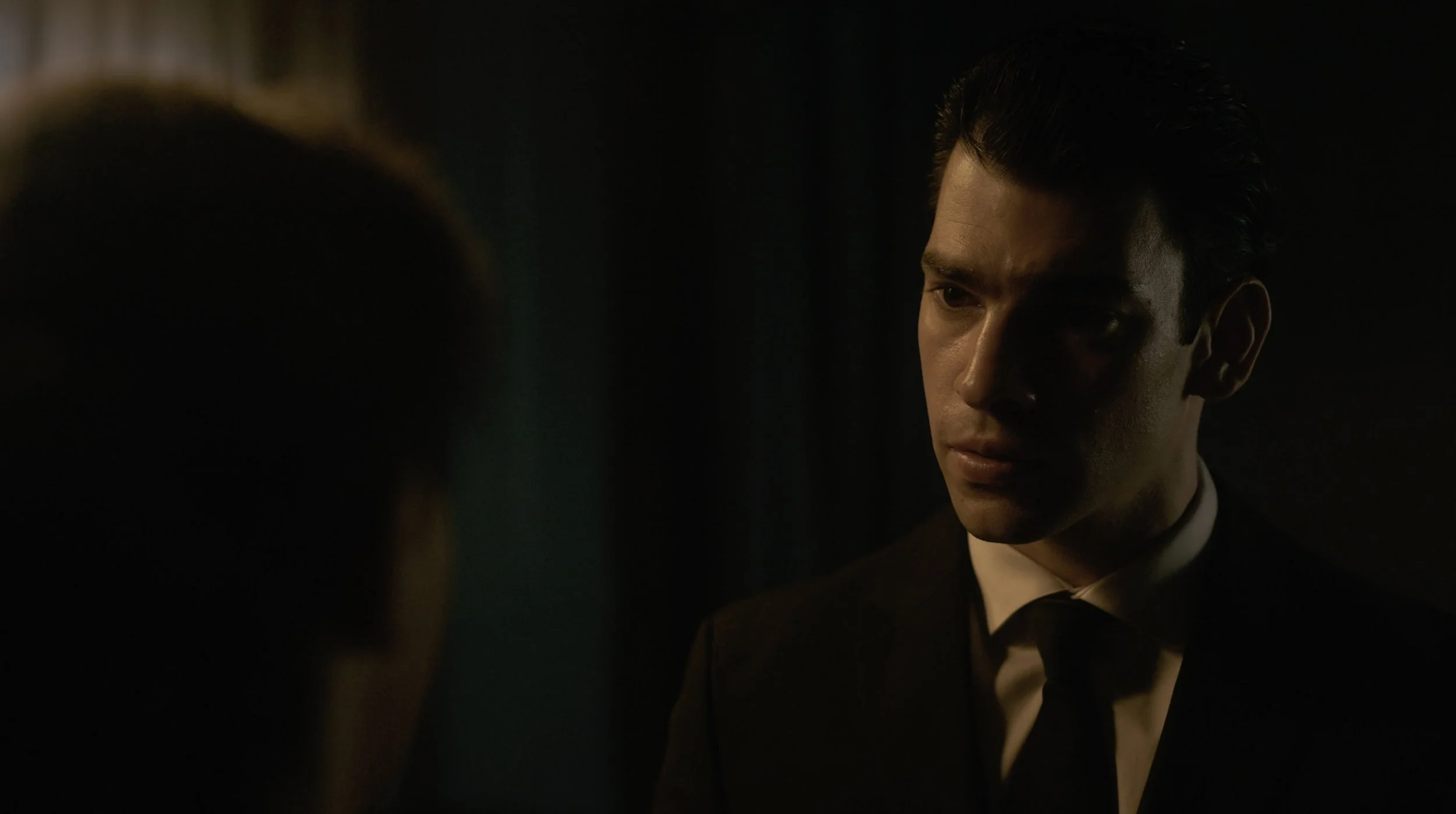 A man in a tuxedo with a white shirt and black bow tie, looking serious, in a dimly lit setting.
