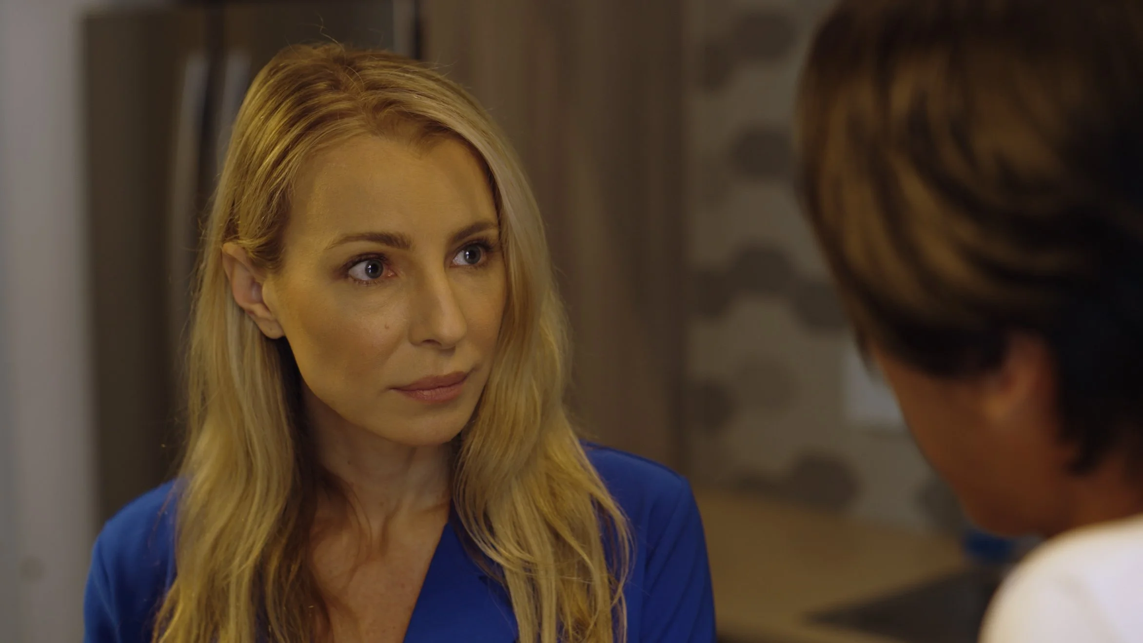 A woman with long blonde hair and blue eyes looking at a person with brown hair, both appearing to be engaged in a conversation indoors.