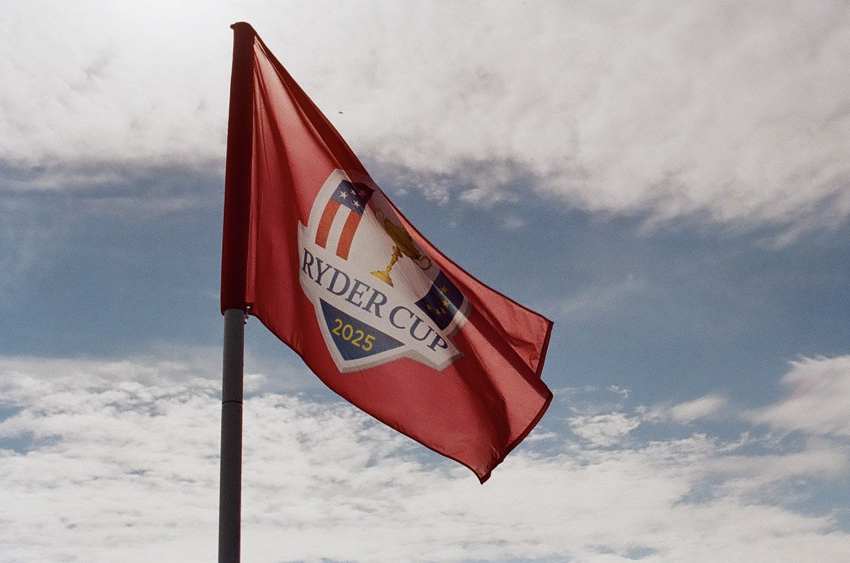 A red flag with the Ryder Cup logo flying in the wind against a partly cloudy sky.