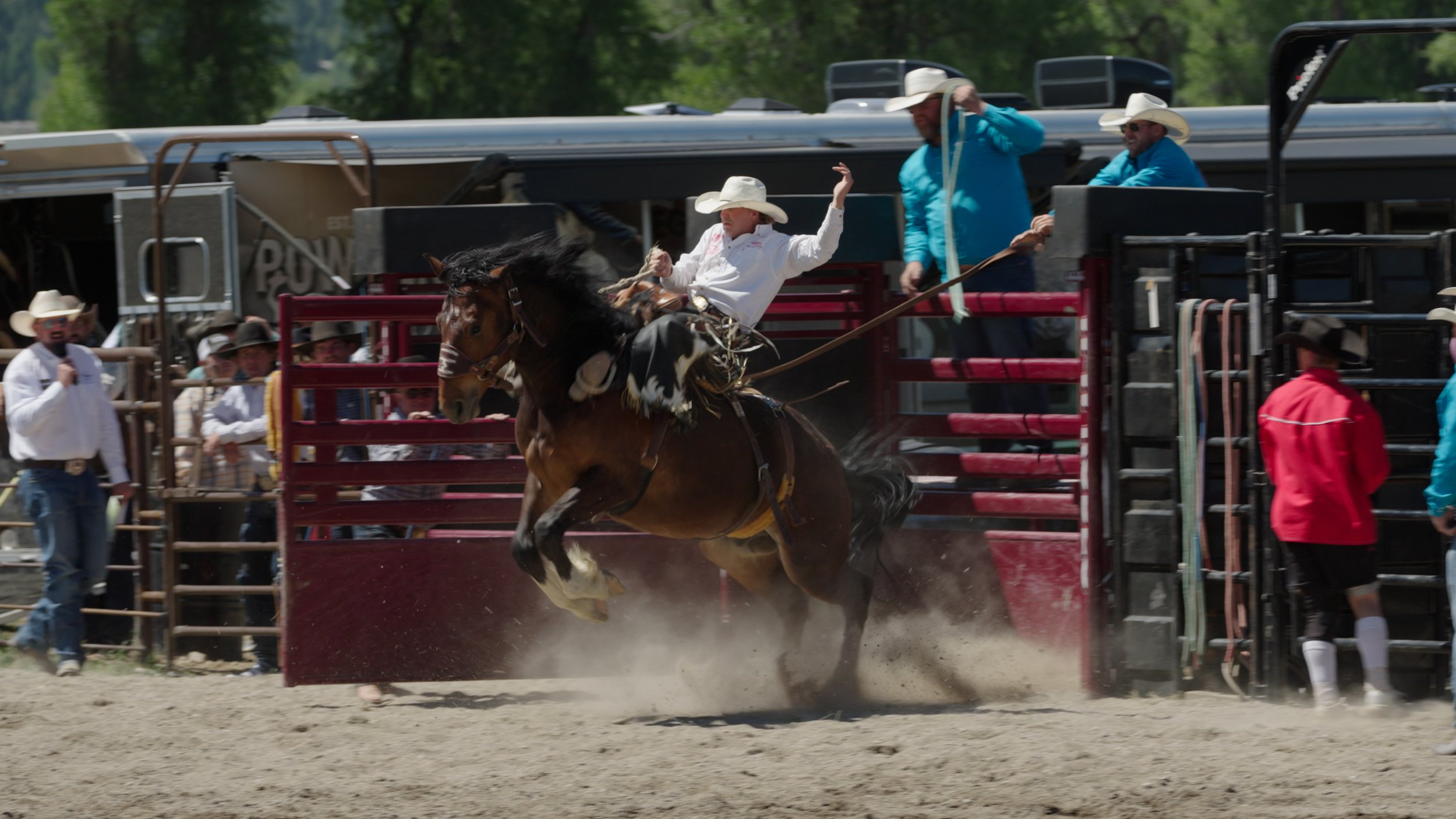 Rodeo cowboy on a bucking bronco in a rodeo arena with spectators and rodeo personnel nearby.