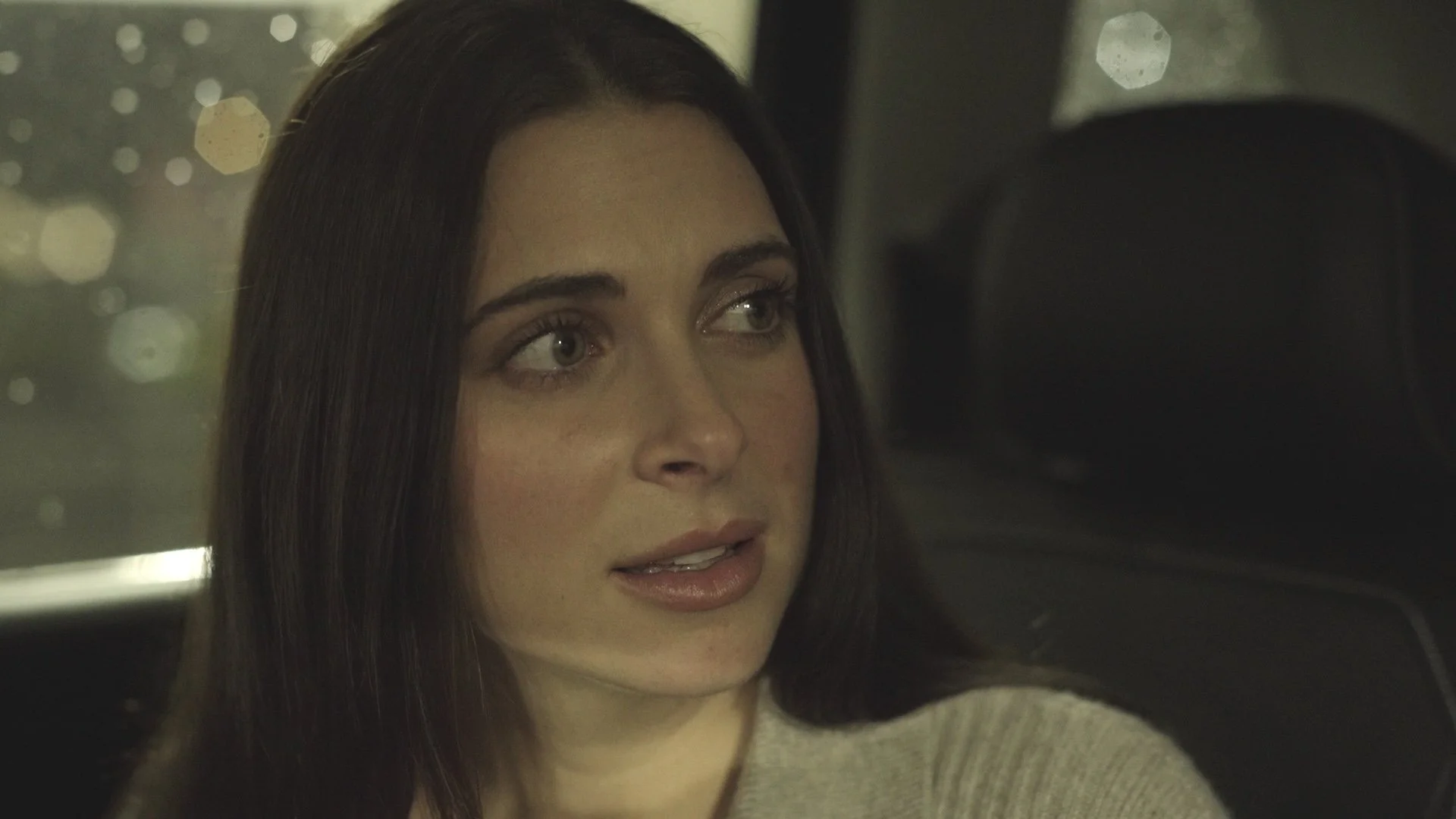 Close-up of a woman with long dark hair and green eyes, looking to her left, with blurred city lights in the background.