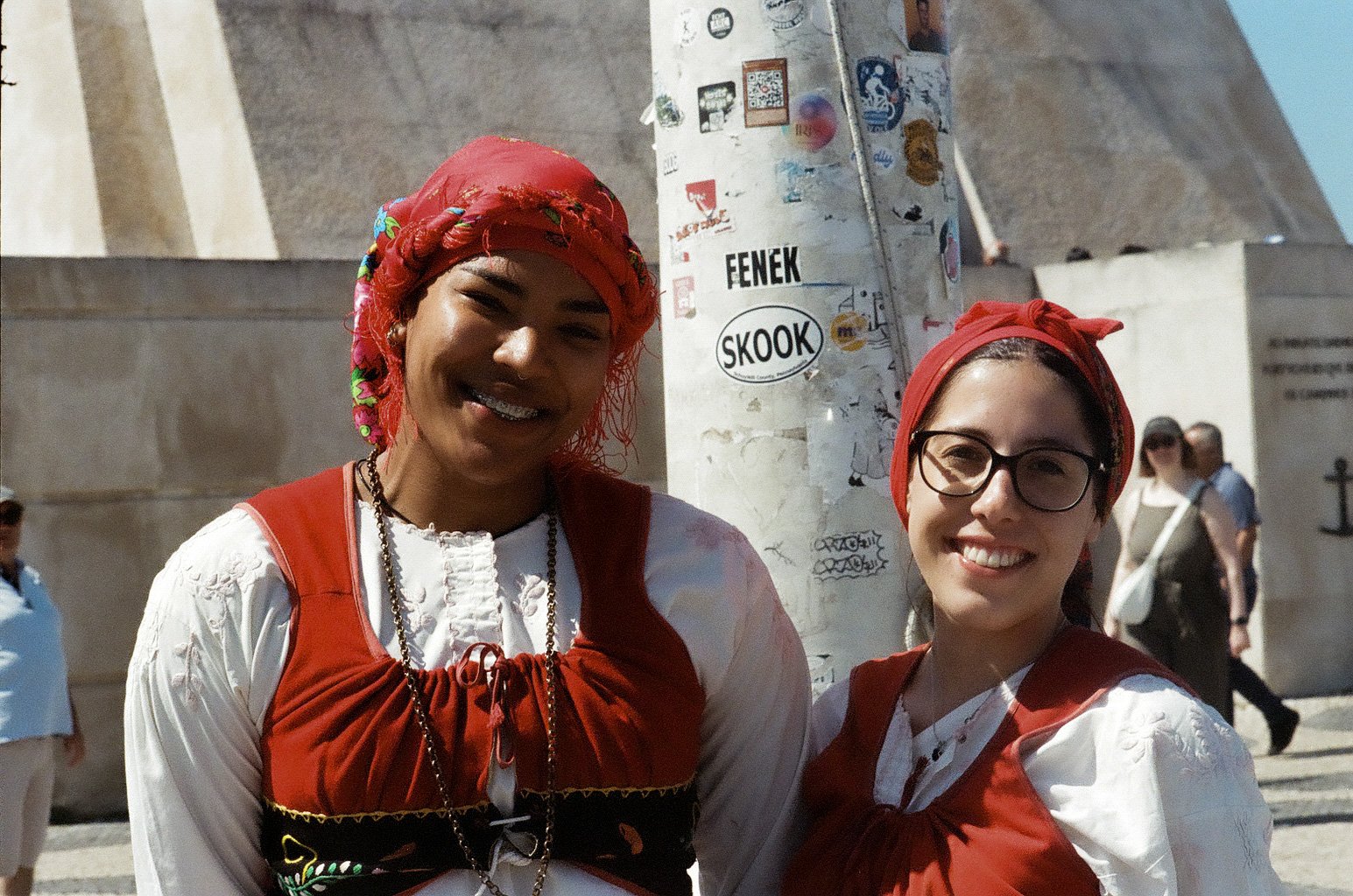 Two women wearing traditional Balkan clothing, one with a red headscarf and the other with a red scarf and glasses, smiling outdoors in front of a pillar covered with stickers and graffiti, with a stone monument and other people in the background.