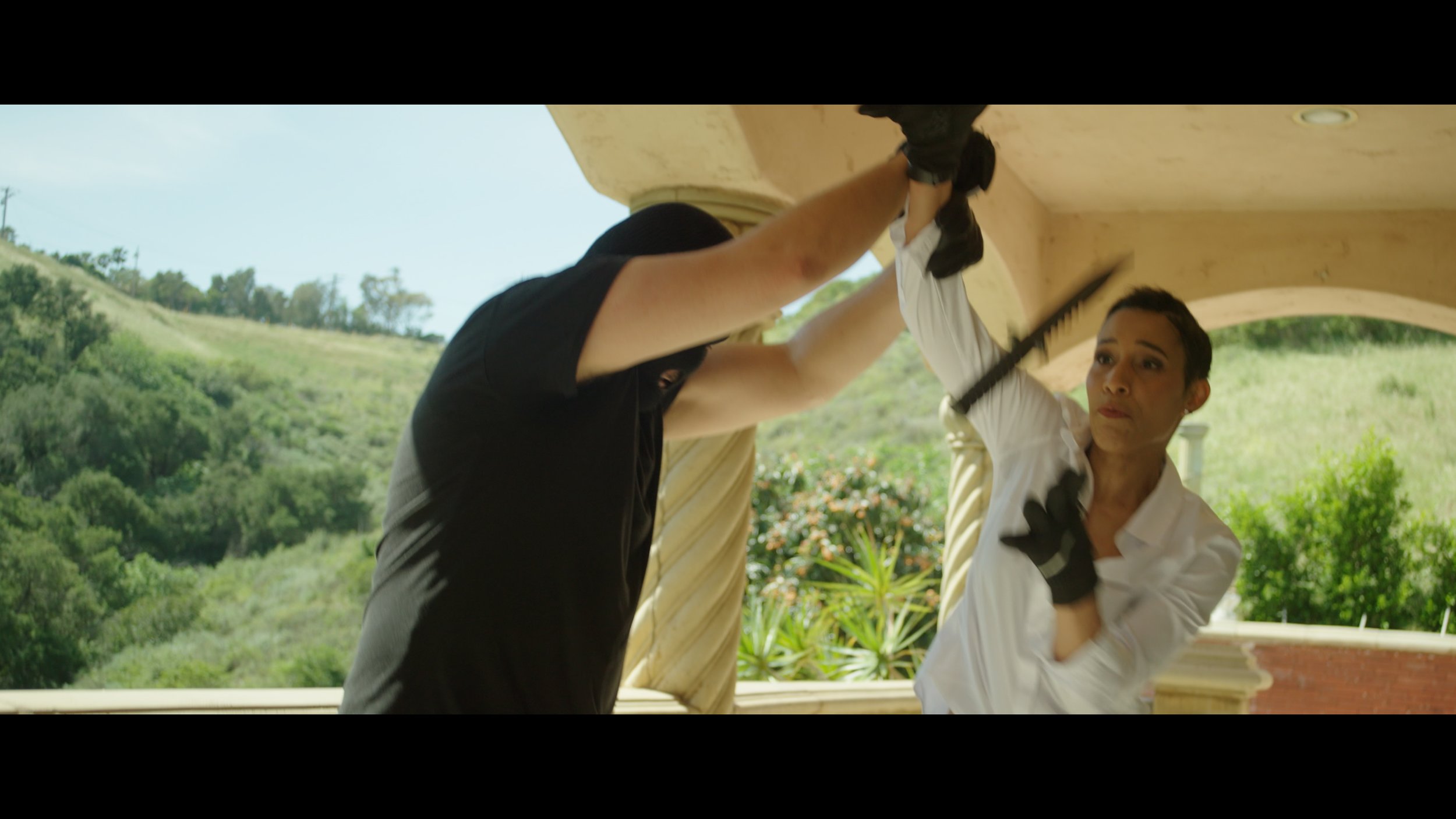 Two people are practicing a martial arts move outdoors beneath an arched structure, with a woman holding a long black stick and looking focused, and a man dressed in black with his face obscured, against a backdrop of green hills and plants.
