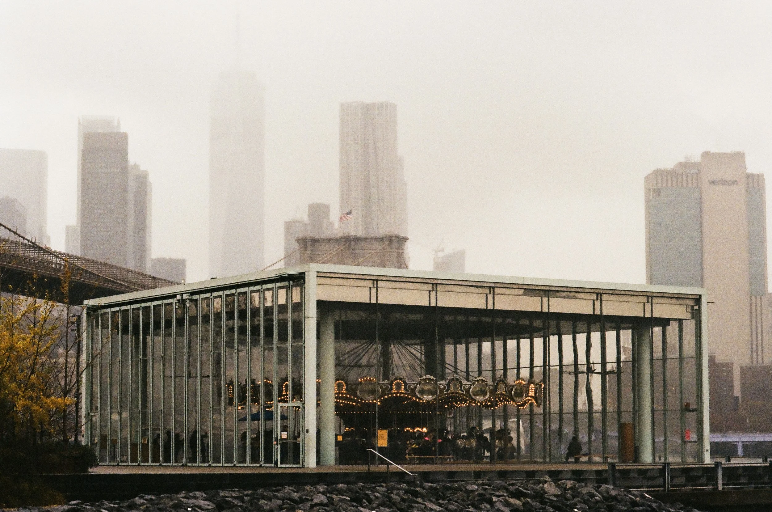 City skyline with tall buildings partially obscured by fog, featuring an American flag on a rooftop, and a modern glass structure with amusement park rides below.