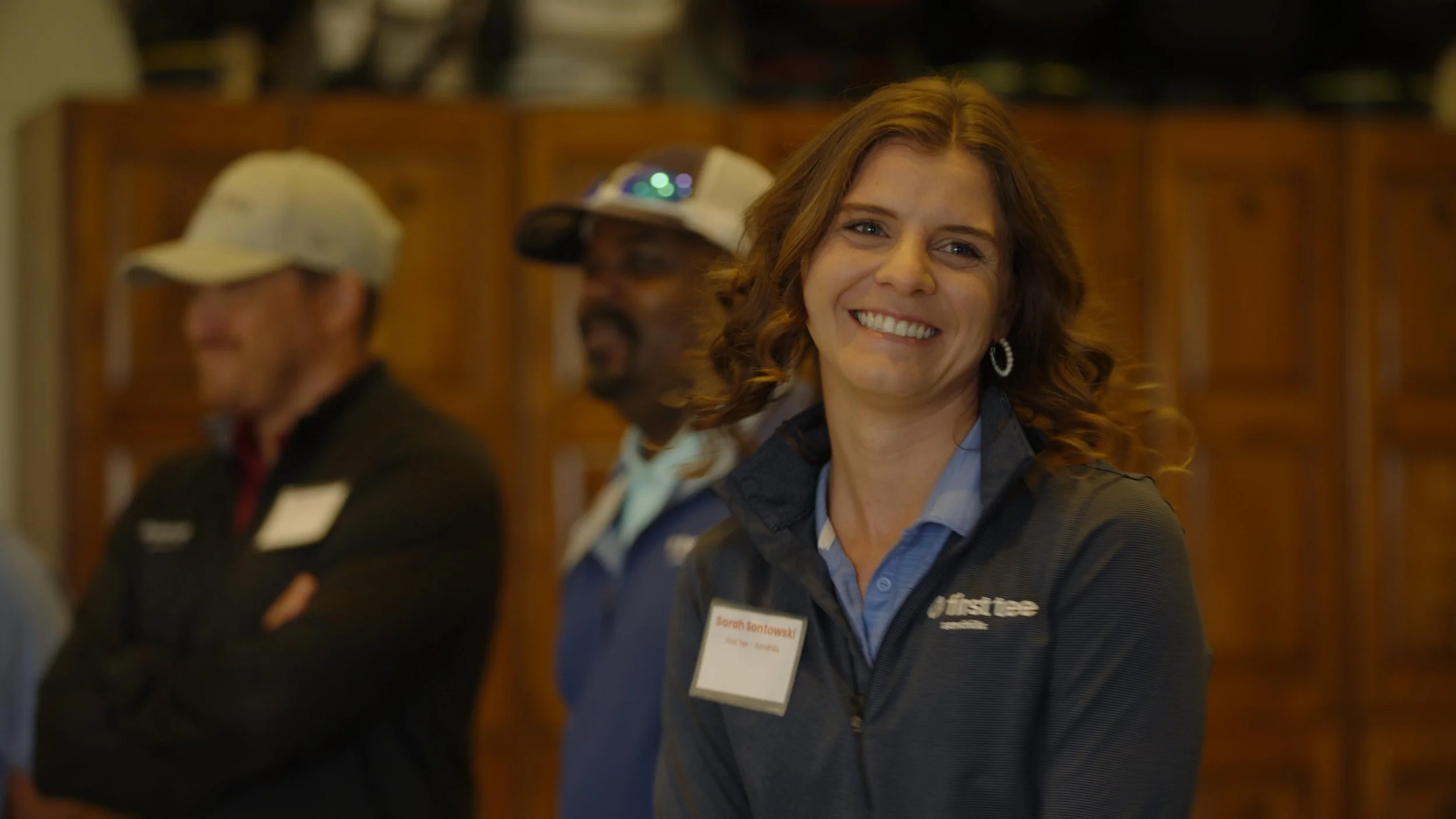 A smiling woman with curly hair wearing a dark jacket and a blue shirt, standing indoors with other people in the background.