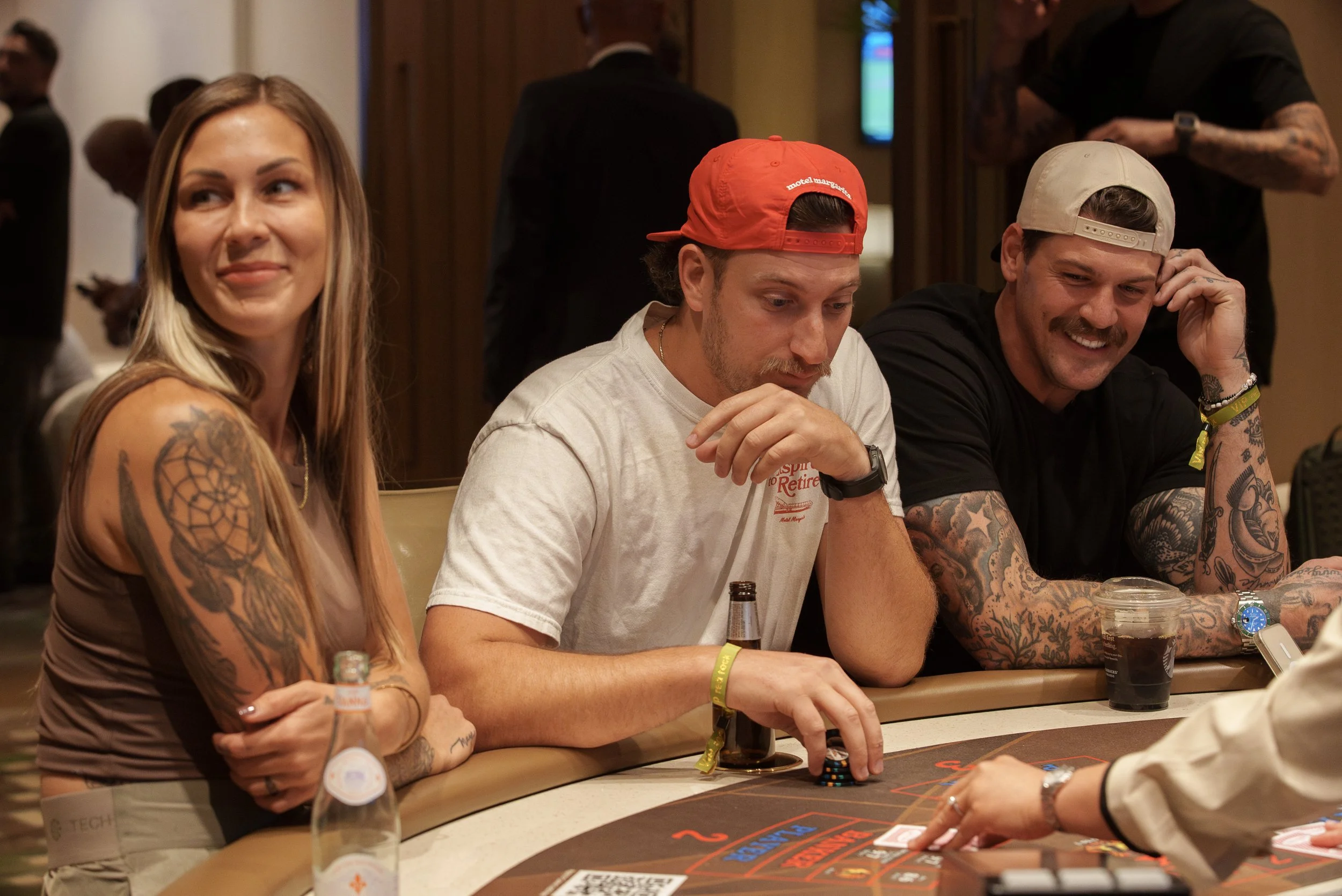 People playing poker at a casino table, with two women and two men, all focused on the game. The women have tattoos and are sitting on each side of the table, while the men are on the opposite side. Drinks and poker chips are on the table.