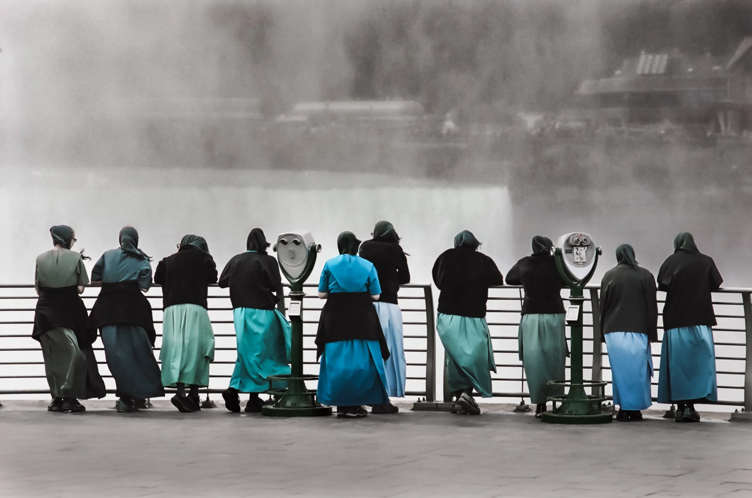 People wearing headscarves and teal skirts standing on a viewing platform, observing a display of the Great Wall of China through a foggy or misty atmosphere.
