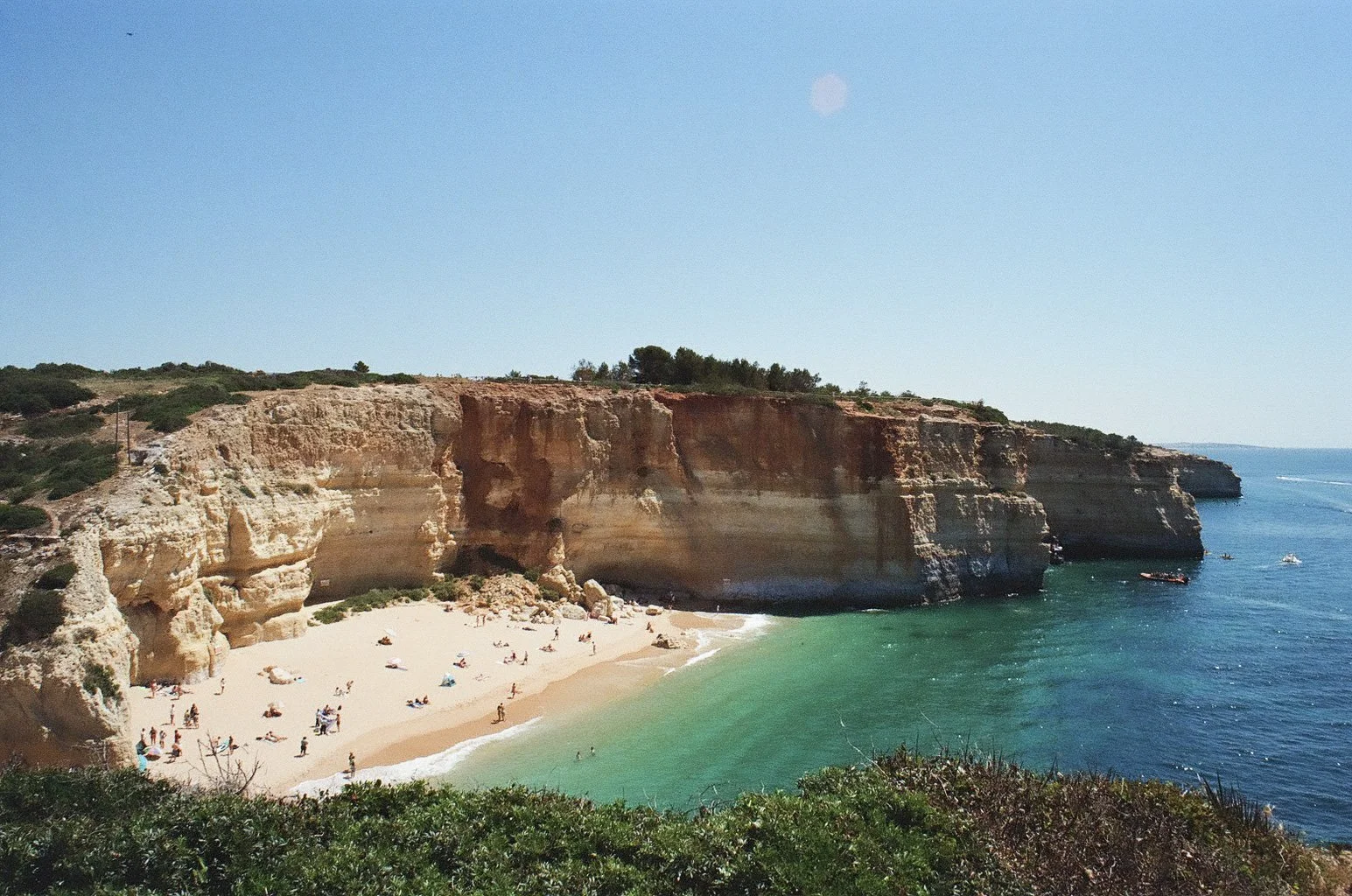Cliffs overlooking a sandy beach with green water, people sunbathing, and boats in the ocean under a clear blue sky.
