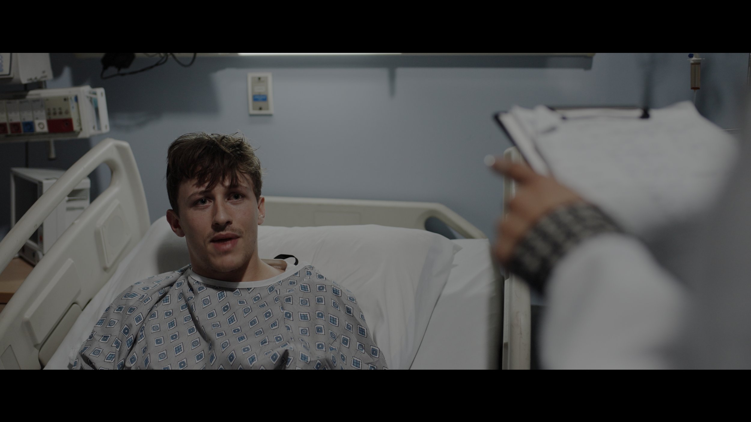 A man in a hospital bed speaking with a healthcare professional holding a clipboard in a hospital room.