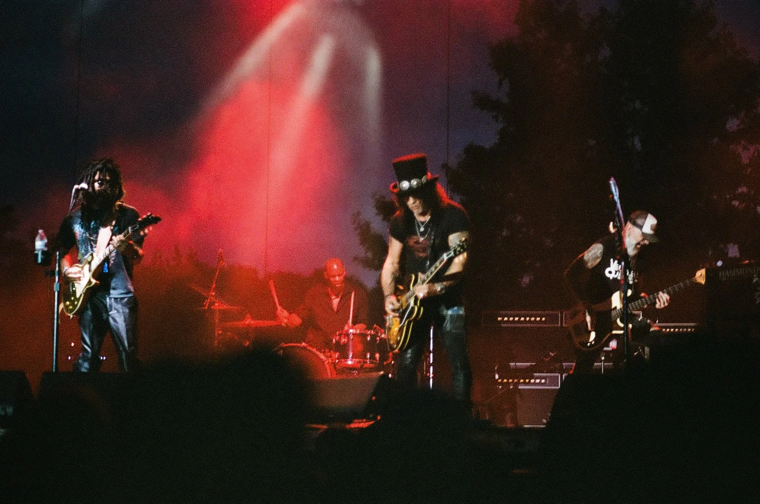 A live outdoor rock concert at dusk with four musicians performing. One is playing an electric guitar, another is on drums, the third on electric guitar wearing a top hat, and the fourth on bass guitar. Stage lights cast a red and purple glow, with t