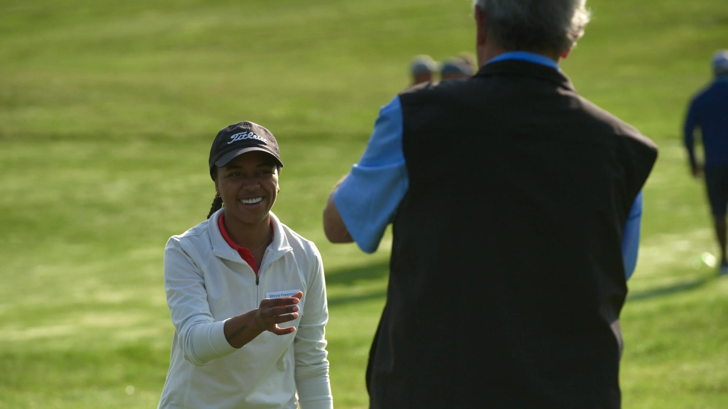 A woman smiling, wearing a golf cap and white jacket, holding a golf tee, talking to a man with gray hair, wearing a black and blue vest, on a grassy golf course.