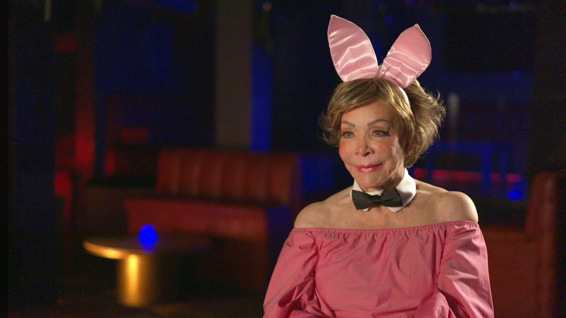 A woman dressed as a bunny in a pink off-the-shoulder top with a black bow tie and pink satin bunny ears, smiling in a dimly lit lounge with red booth seating and blue accent lighting.