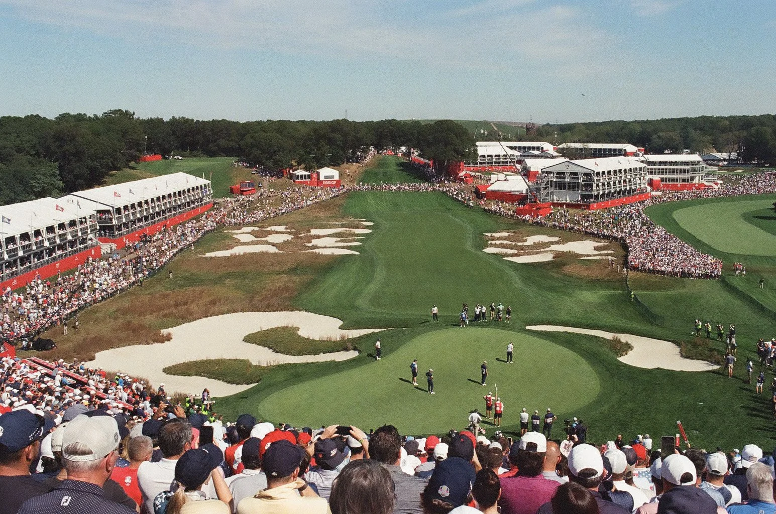Crowd watching a professional golf tournament at a large outdoor golf course with multiple grandstands and spectators along the fairway and around the green.