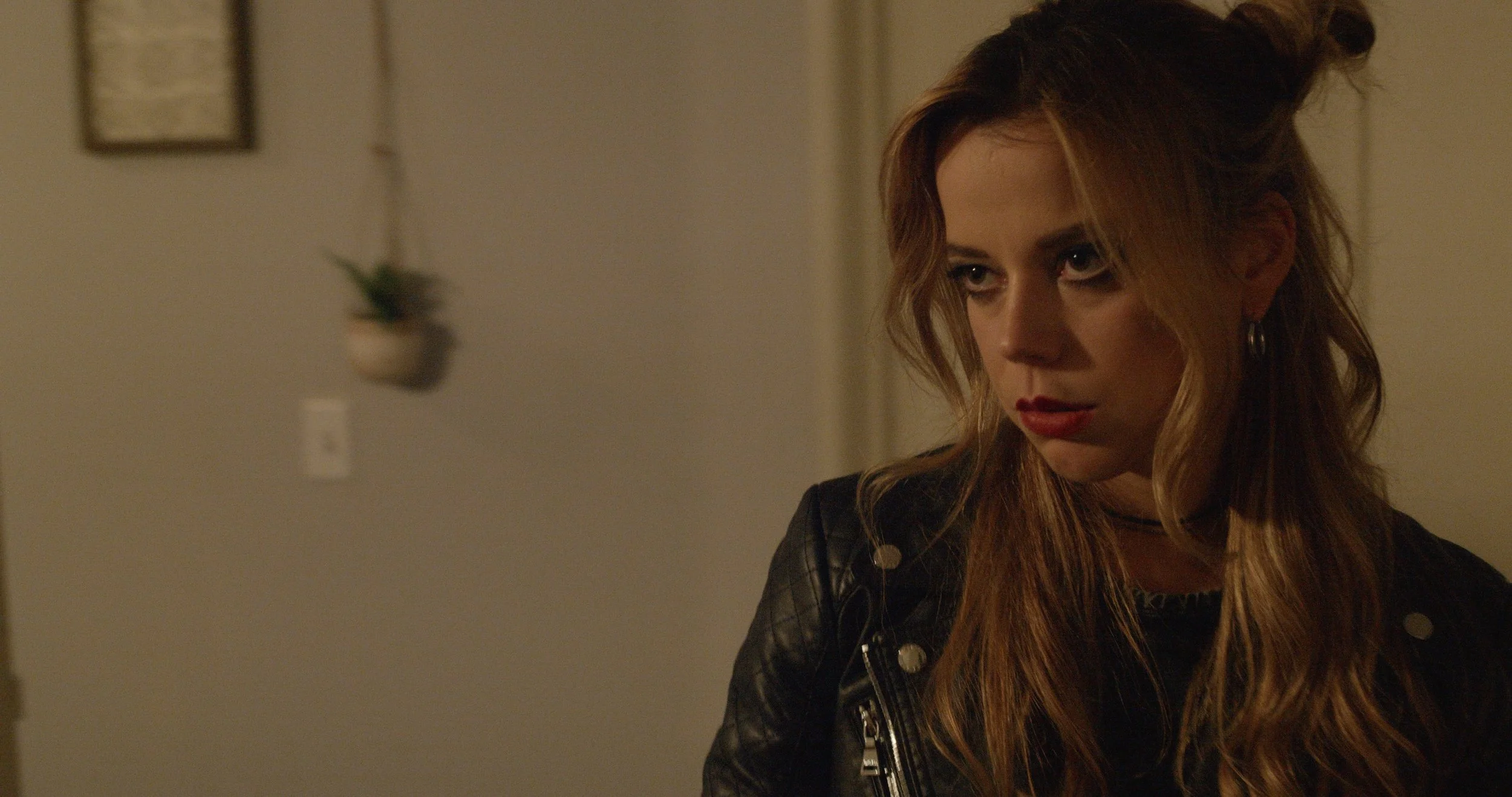 A woman with long wavy hair, wearing a black leather jacket, looking downward in a dimly lit room.