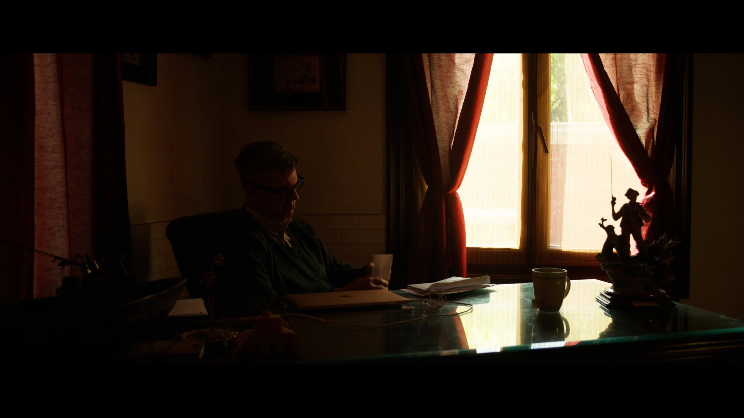 A man sitting at a dining table reading a paper, with decorative figurines on the table, behind a window with partially open red curtains, illuminated by natural sunlight.