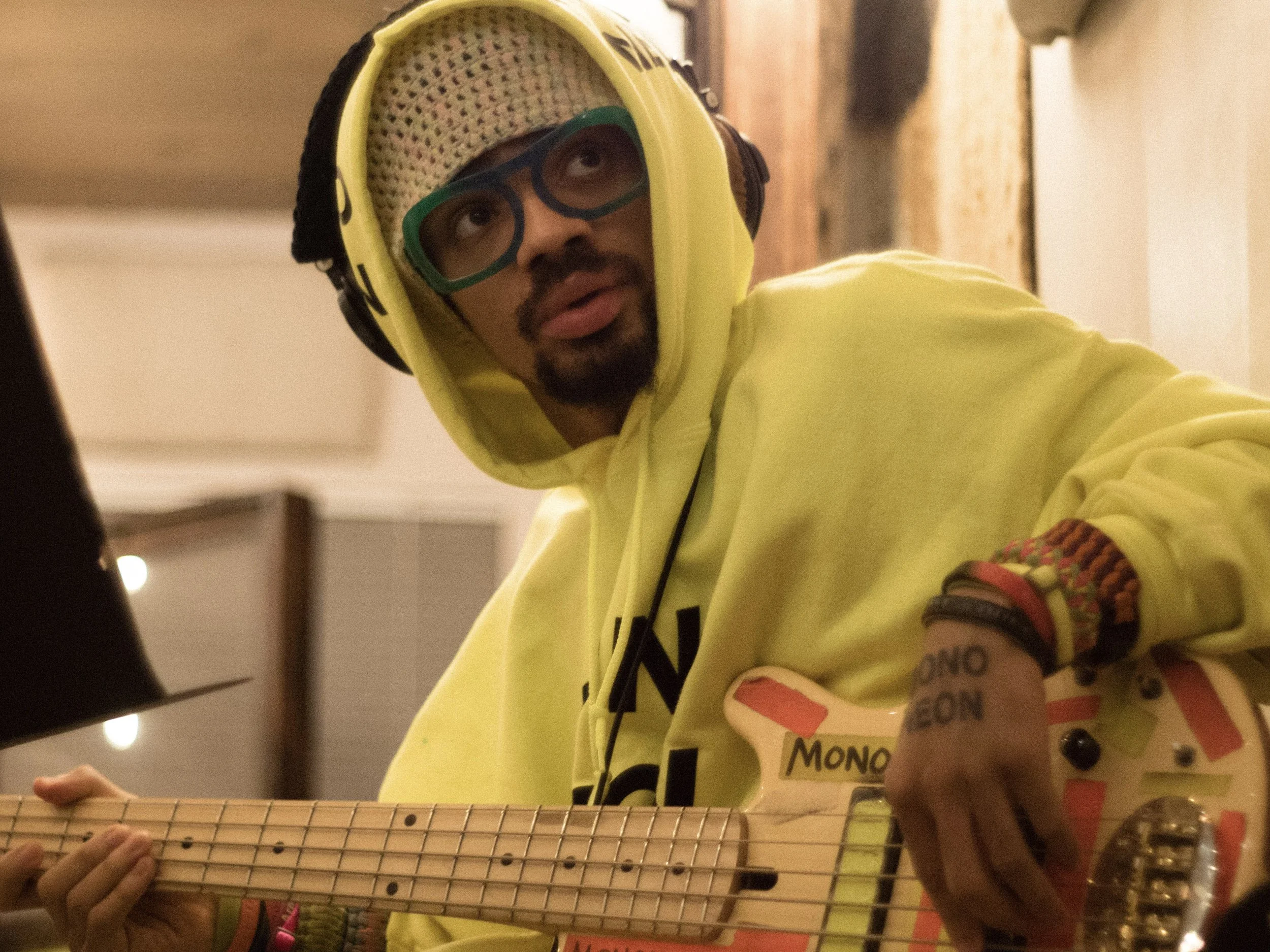 Person wearing yellow hoodie, green glasses, and hat with colorful mesh pattern, holding a guitar in a room with warm lighting.