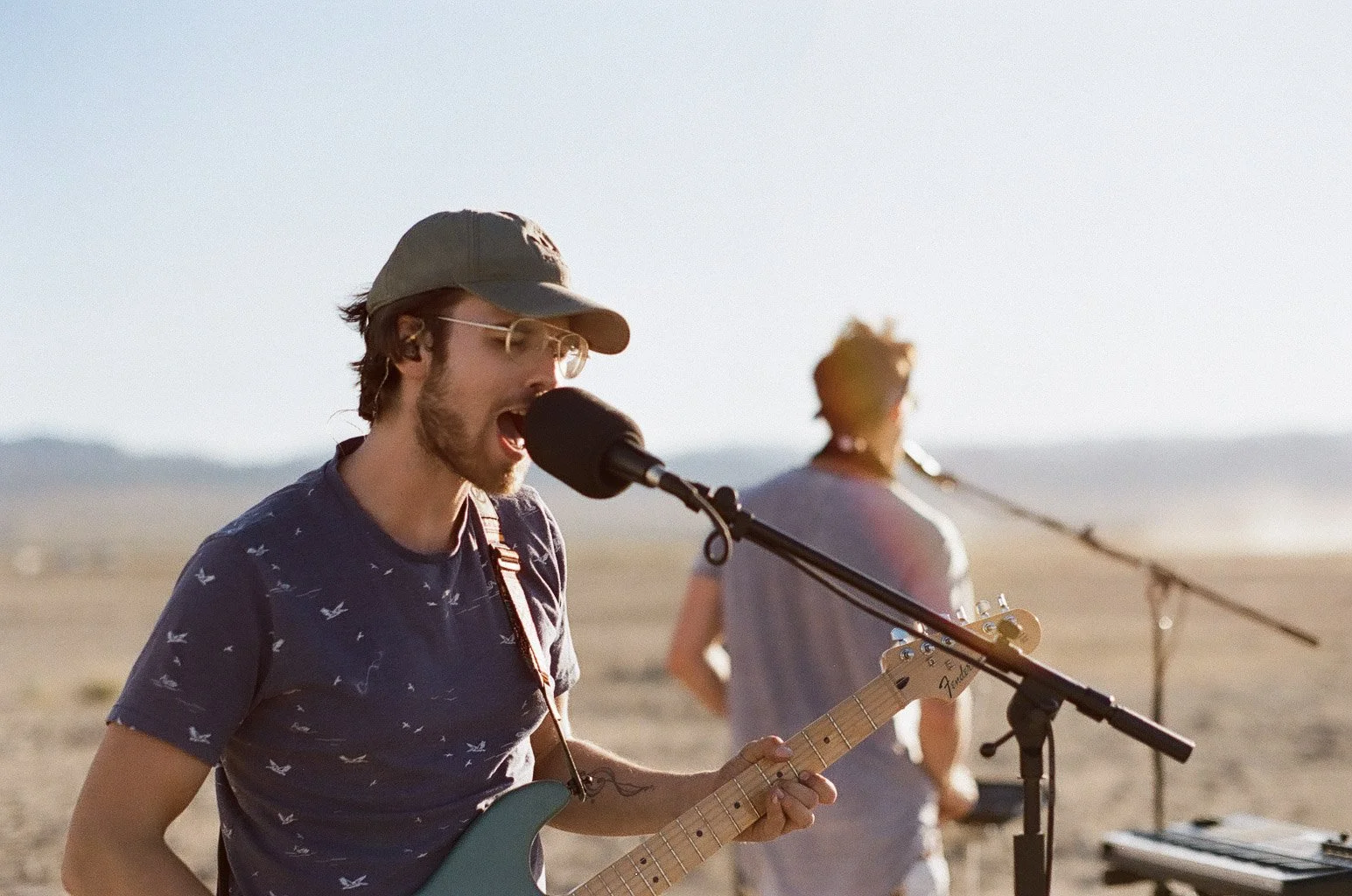 A musician singing into a microphone while playing an electric guitar in a desert landscape with another person in the background near a microphone stand.