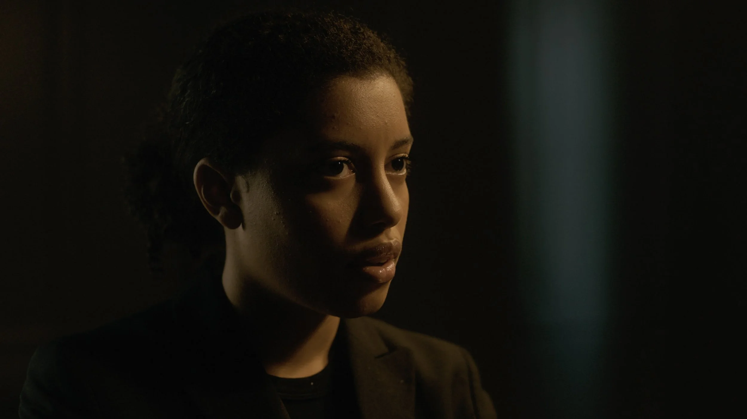 Close-up of a young woman with natural curly hair and medium skin tone, looking thoughtfully into the distance in low lighting.