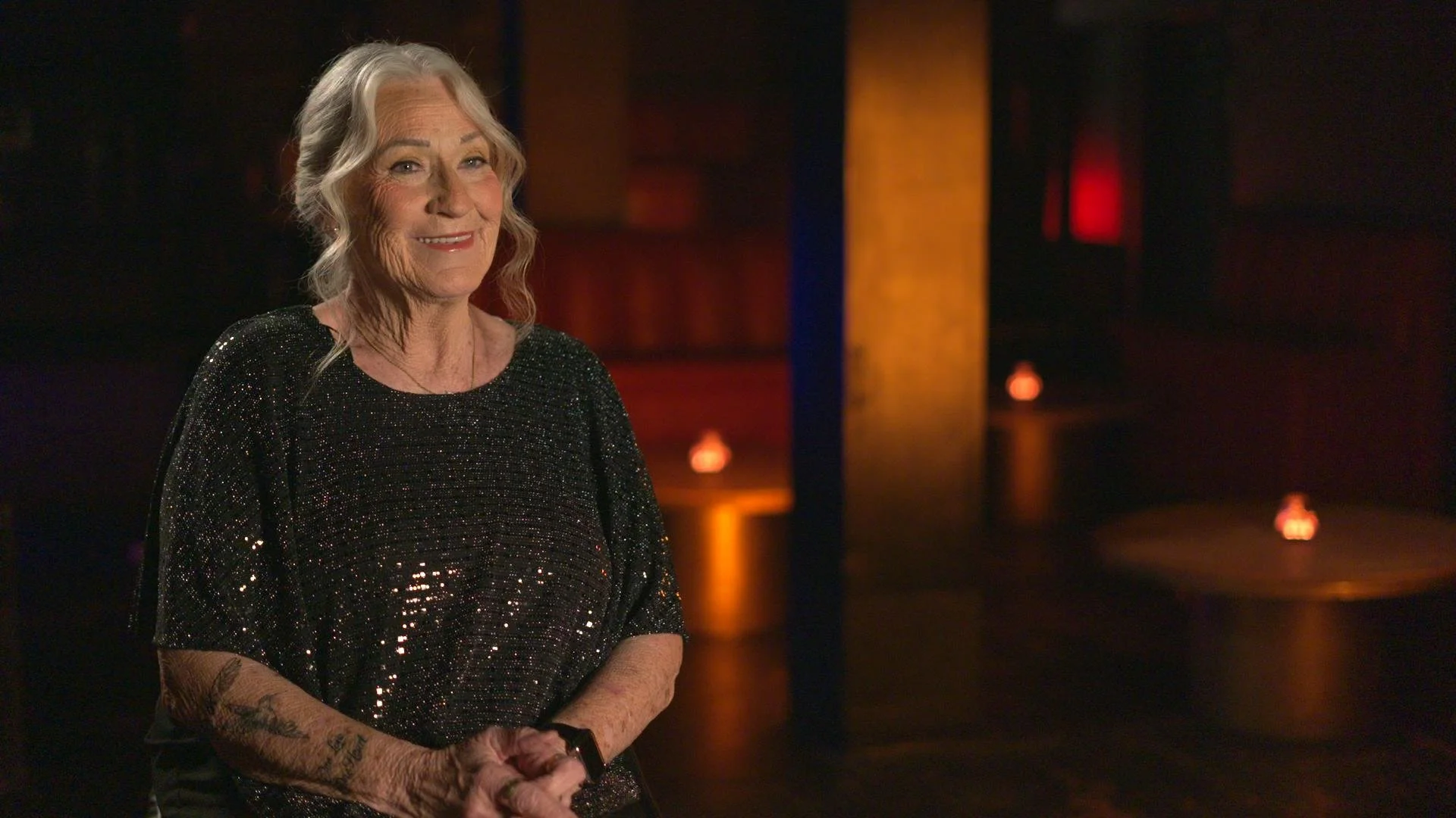 An elderly woman with gray hair and tattoos on her arms, smiling and wearing a black sparkly top, standing in a dimly lit room with warm orange and red lighting.