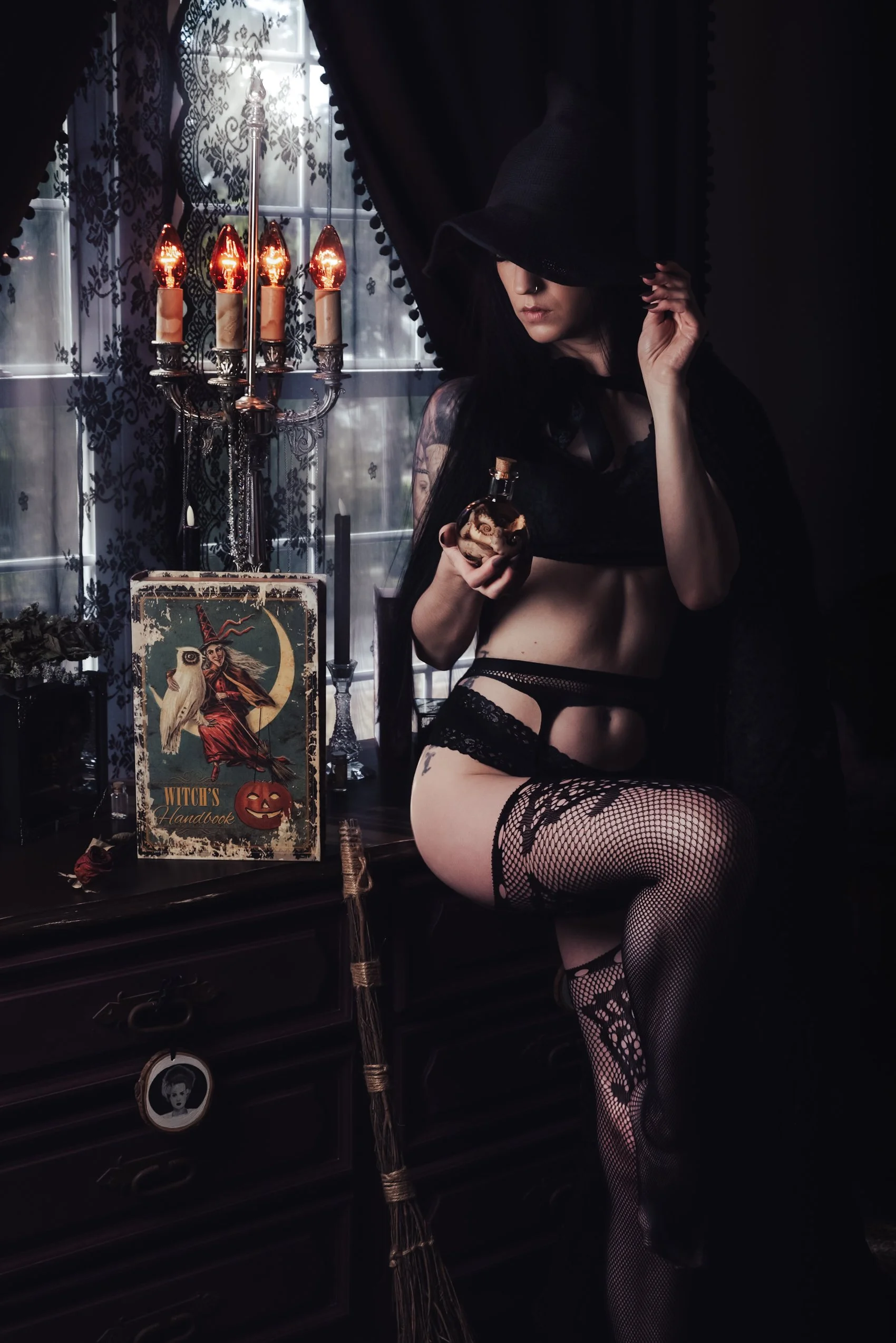 A woman dressed in black lingerie and fishnet stockings, covering her eyes with a black hat, holding a small bottle, sitting beside a vintage dresser decorated for Halloween with creepy posters, a candlestick, and candles, inside a dimly lit room wit