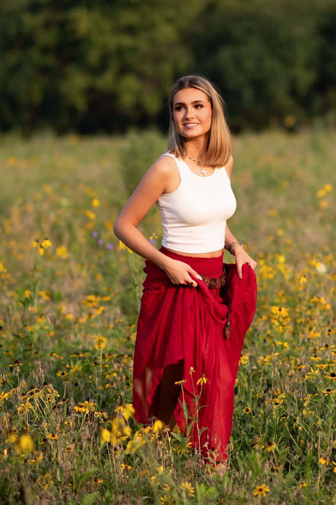 A young woman in a white tank top and a flowing red skirt stands in a field of yellow flowers with green trees in the background, smiling and looking into the distance during sunset.