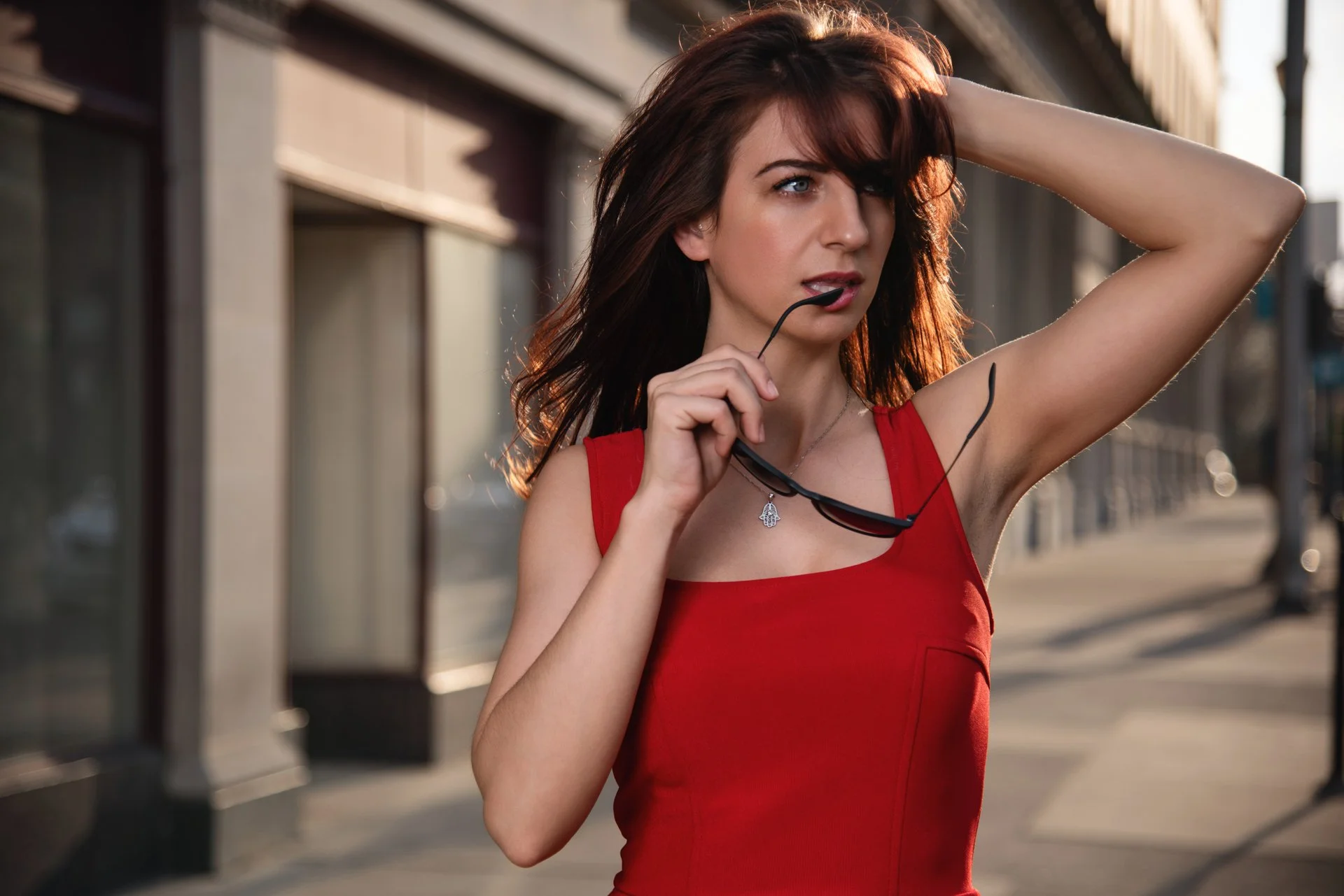 A woman with red hair wearing a red sleeveless top is outdoors on a sidewalk, holding sunglasses near her mouth, with one hand on her head, looking contemplative.
