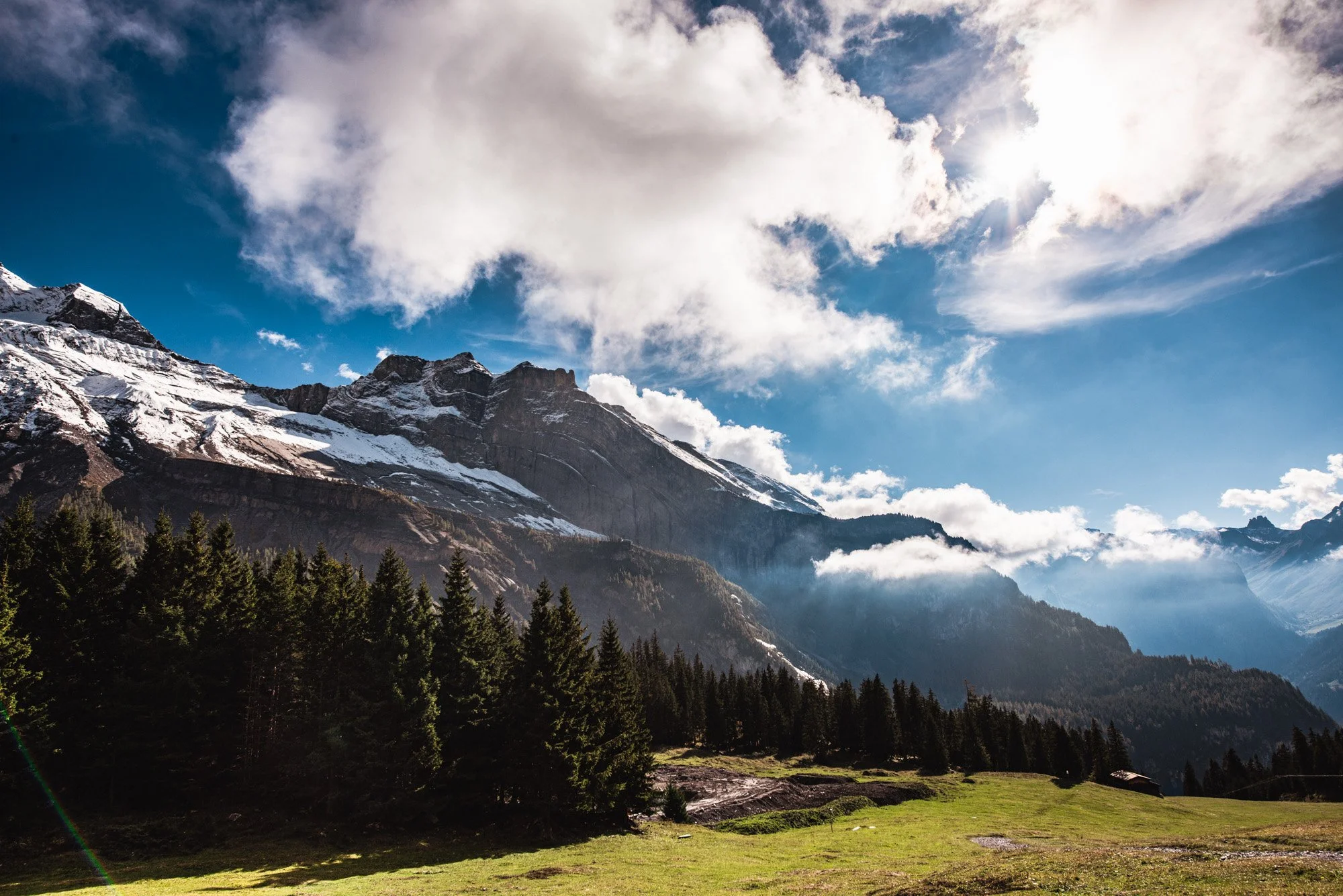 Snow-capped mountain peaks with rocky cliffs under a partly cloudy blue sky with the sun shining through clouds and a lush green forest at the base.