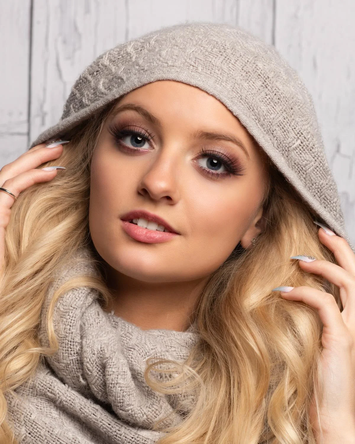A young woman with long blonde hair wearing a beige knit hat and scarf, touching her hair with both hands, against a white rustic background.